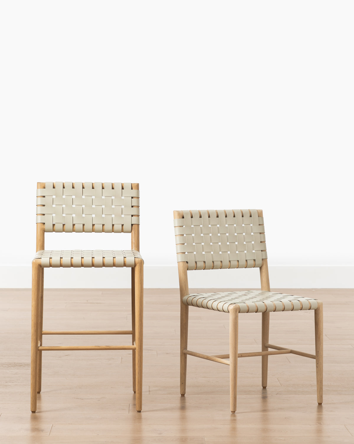Two Greely Dining Chairs with light woven seats and backs are side by side on a light wood floor against a white background; the left chair is barstool height, while the right is standard height, by McGee & Co.