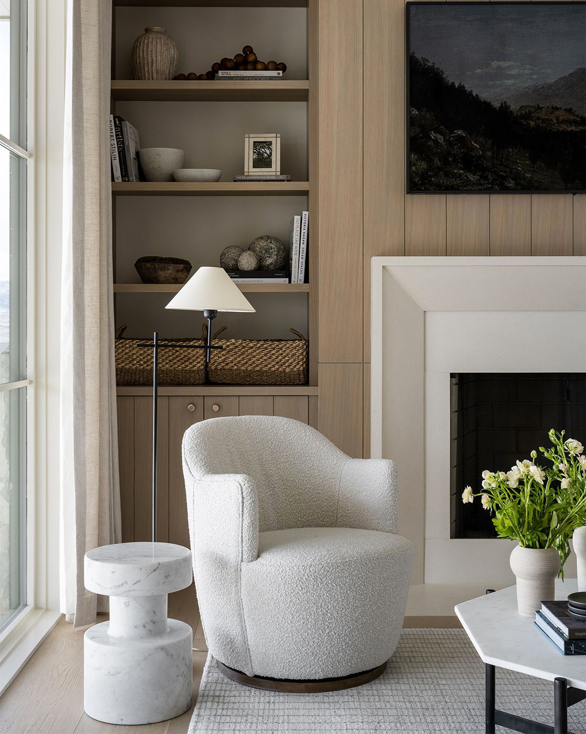 A cozy, modern living room corner features the Gulliver Swivel Chair in soft boucle, a round marble side table with a flower vase, a standing lamp, built-in wooden shelves with decor, and a white fireplace.