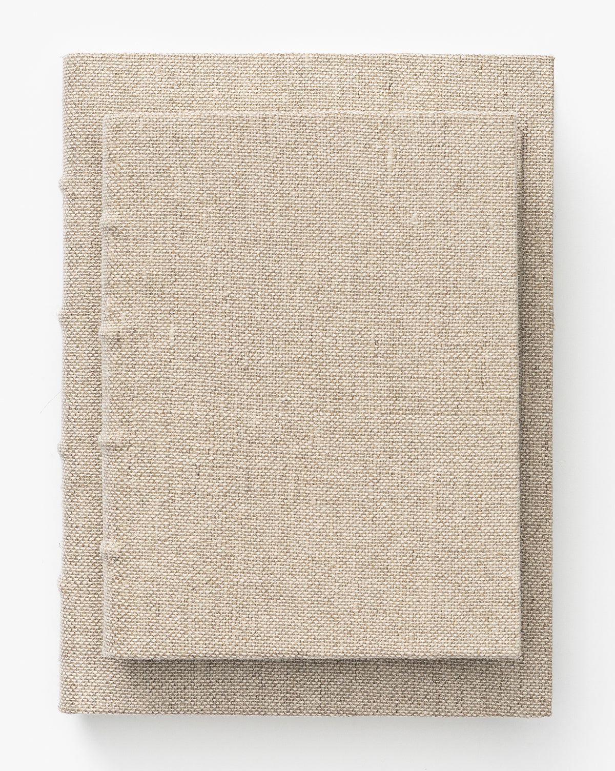 Two beige Handcrafted Linen Books with linen fabric hardcovers are stacked on a white surface. The smaller book rests neatly atop the larger one, both featuring traditional binding and plain covers.