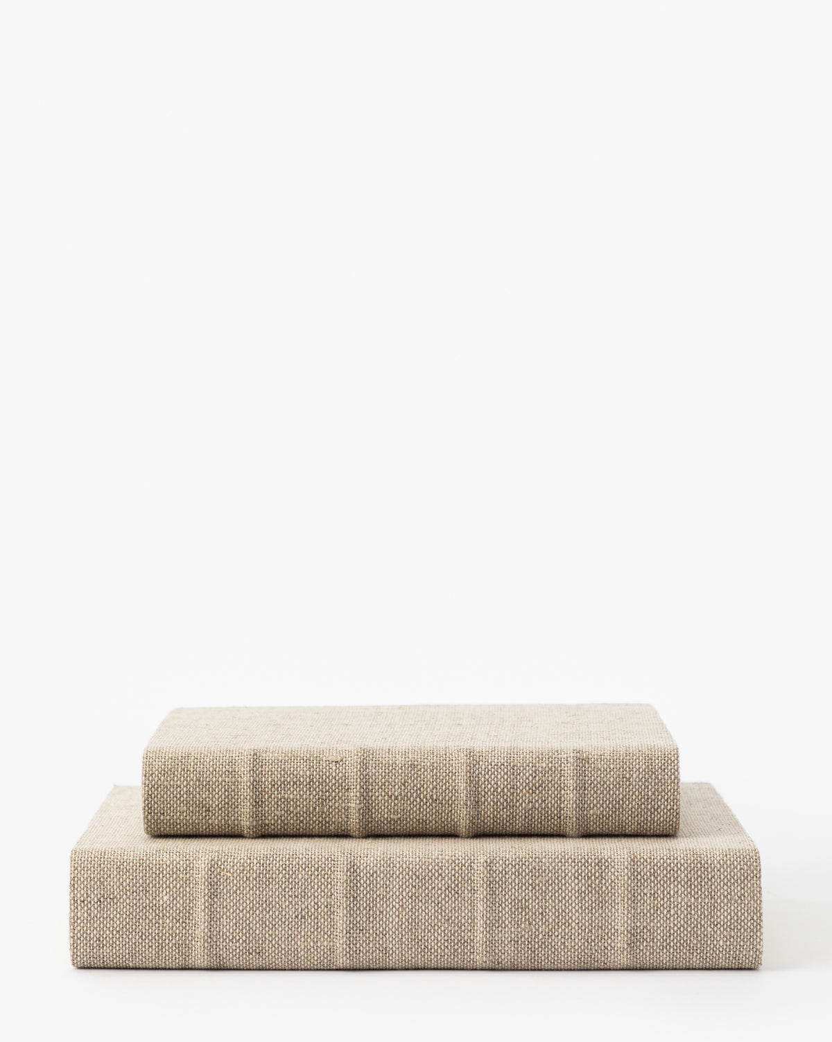 Two Handcrafted Linen Books in beige with textured covers are stacked on a white surface against a plain background. The top book is smaller, and both feature traditional hardcover binding.
