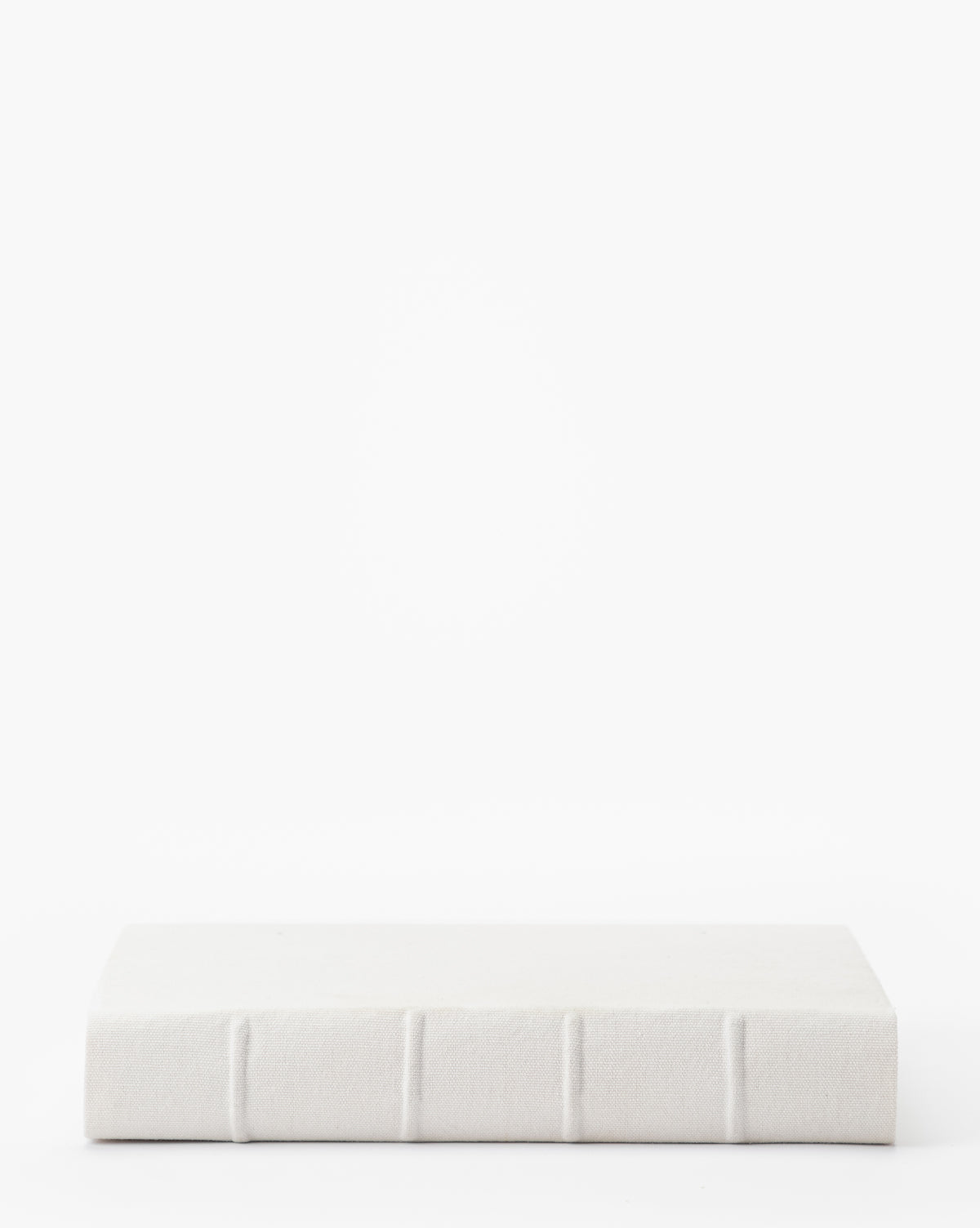 A closed Handcrafted Linen Book with a textured white linen cover and visible spine ridges rests on a white surface in front of a white background.