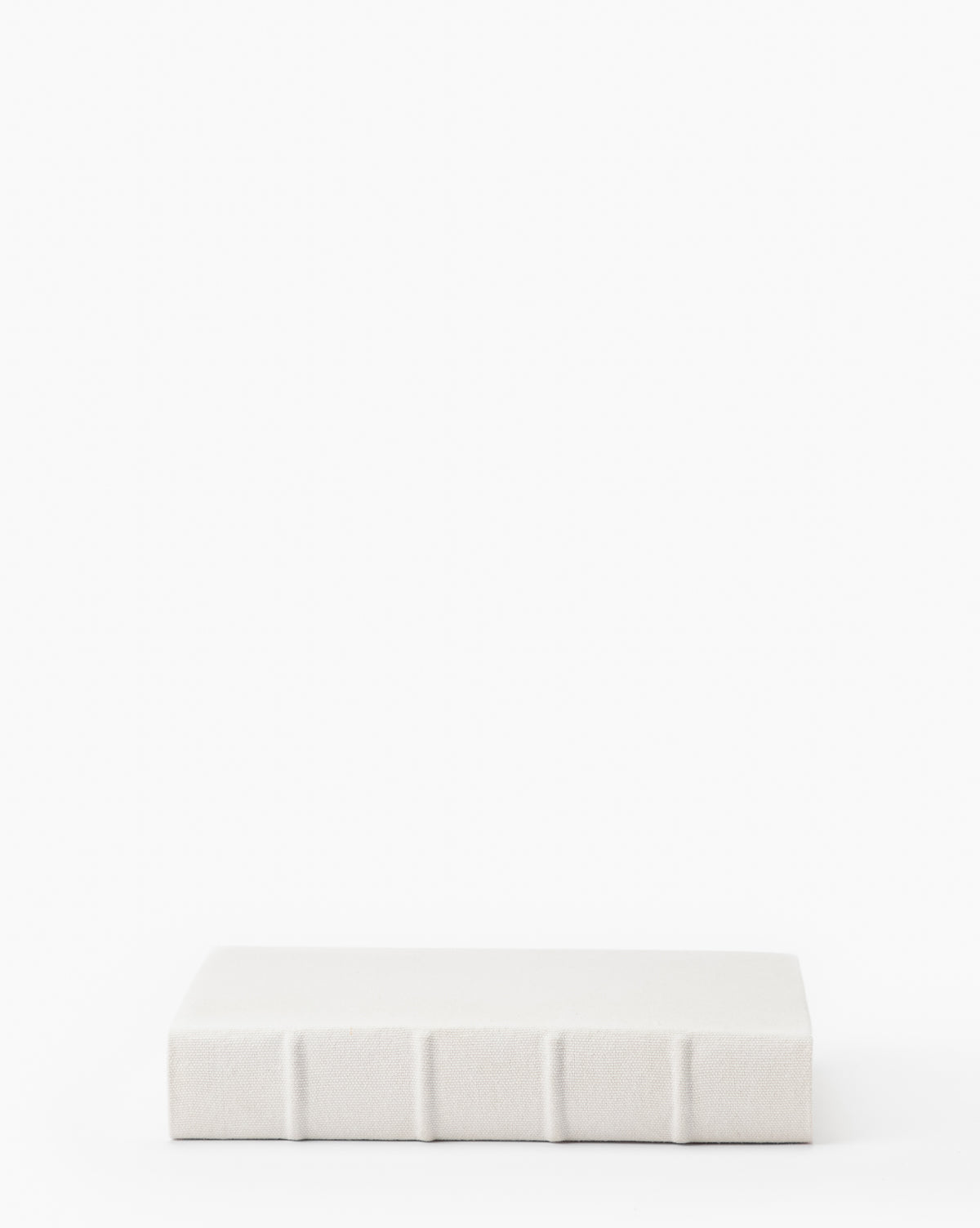 A closed Handcrafted Linen Book with a plain white linen cover and visible spine ridges rests on a white surface, highlighting its traditional binding craftsmanship.