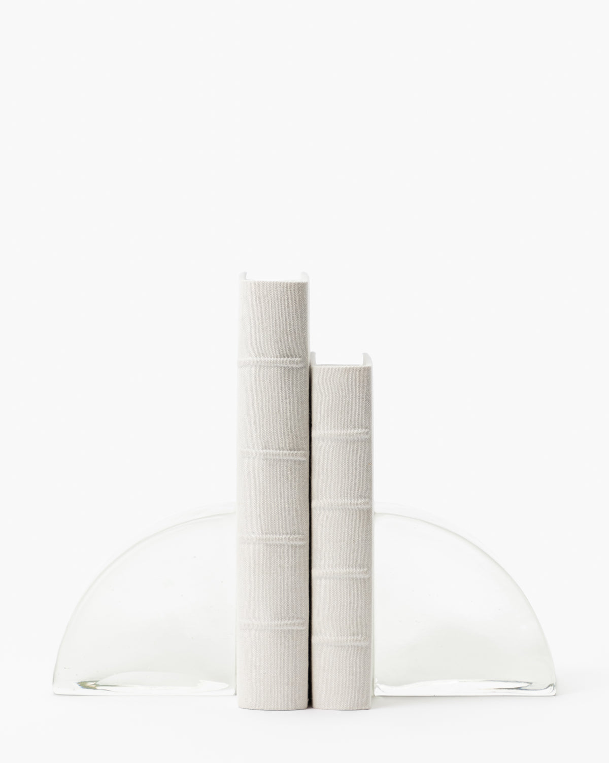 Two Handcrafted Linen Books with traditional binding stand upright between two clear, semi-circular glass bookends on a white background, showcasing a minimalist scene with no visible text on the spines.