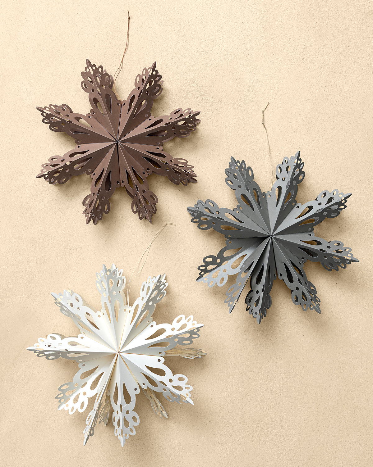 The Handmade Assorted Recycled Paper Snowflakes (Set of 3) features ornaments in brown, gray, and white with intricate cut-out details and strings for easy holiday decorating, shown hanging against a plain beige background, by McGee & Co.