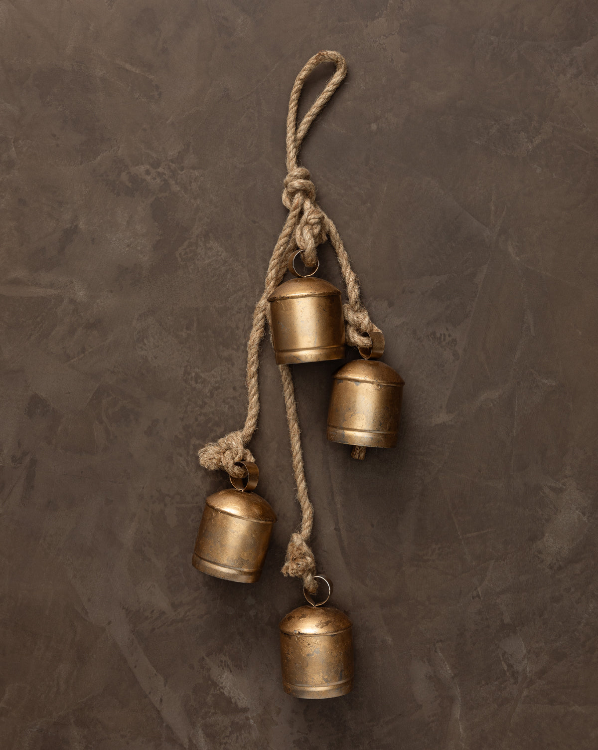 The Hanging Iron Bells set features four small, gold-finished bells with rounded tops hanging at varying lengths from a twisted rope—ideal for holiday décor against a neutral backdrop, by McGee & Co.