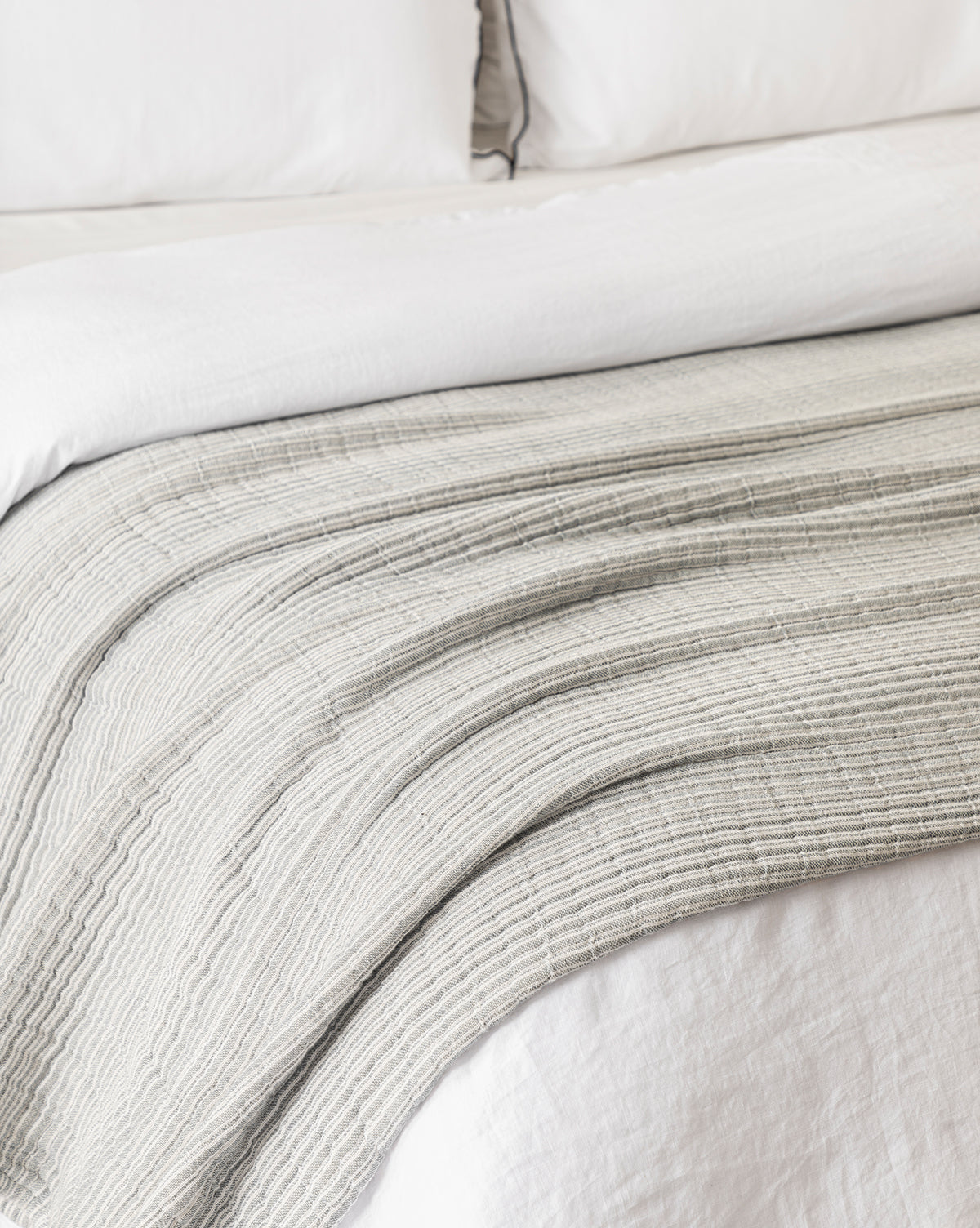 A neatly made bed features the Hanson Striped Cotton Coverlet in light gray draped smoothly at the foot, creating a soft, cozy, and inviting look that highlights timeless and neutral bedding styles.