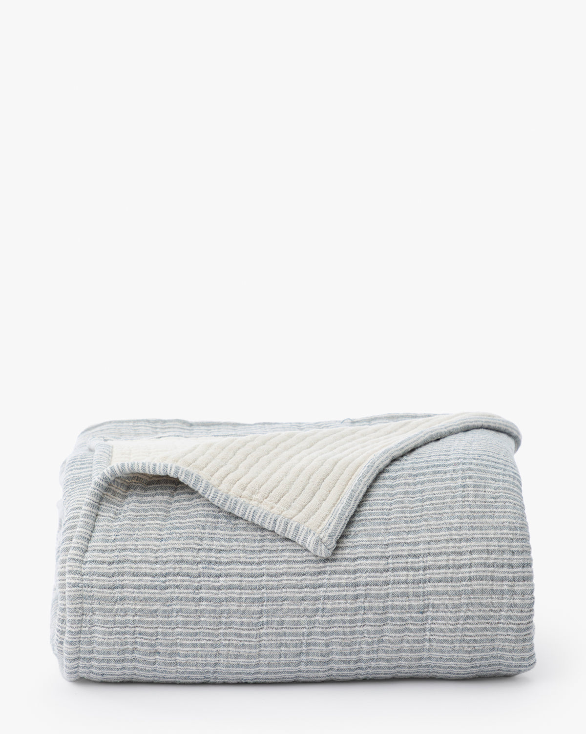 The Hanson Striped Cotton Coverlet, in light blue with textured stripes and a cream inner lining, is neatly folded with a corner flipped over. Set on a white background, it’s versatile neutral bedding perfect for any style.
