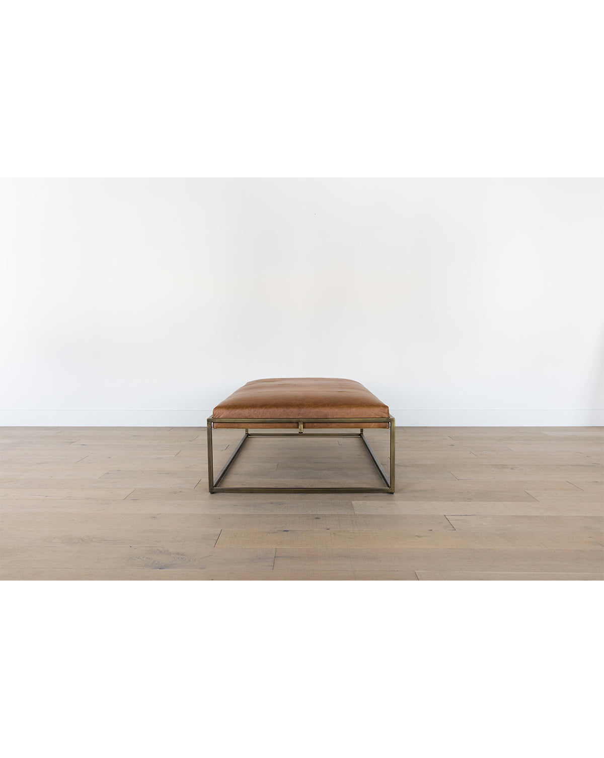 The Harlow Leather Coffee Table, featuring a sleek brass-finished frame and aniline-dyed leather top, sits on a light wooden floor against a plain white wall, by McGee & Co.