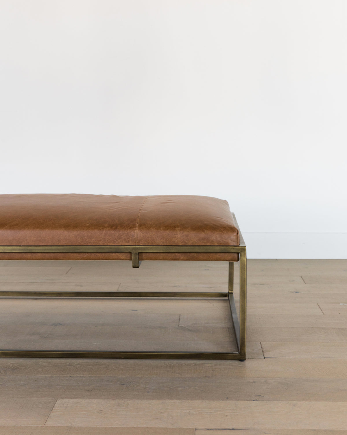 The Harlow Leather Coffee Table, featuring an aniline-dyed leather cushion and a sleek brass-finished frame, sits on a light wooden floor against a white wall. Only the right half of the table is visible, by McGee & Co.