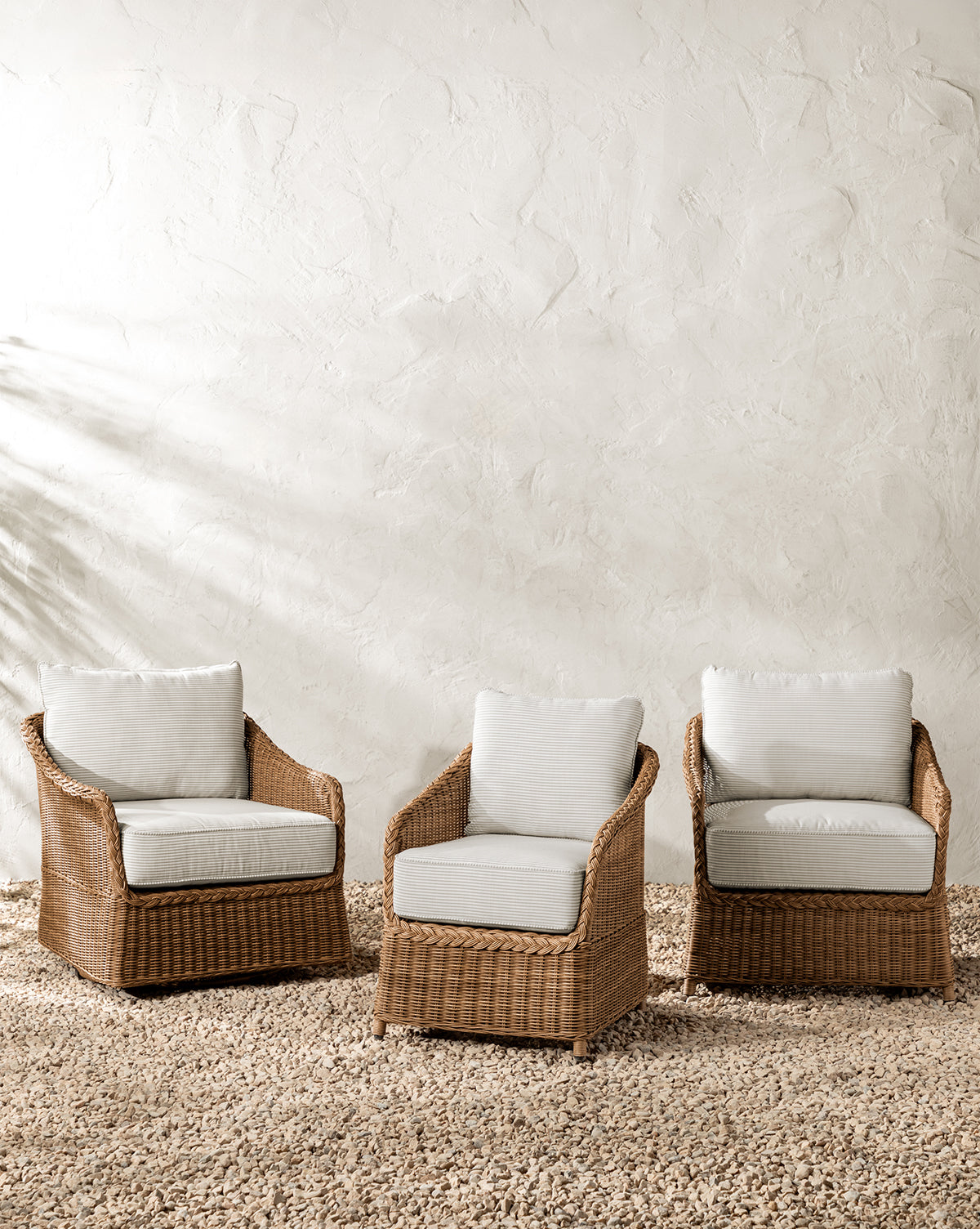 Three Haviland Outdoor Lounge Chairs with Perennials Taupe Stripe cushions are arranged on a pebble floor against a textured white wall. Soft natural light casts gentle shadows, creating a calm and inviting atmosphere by McGee & Co.