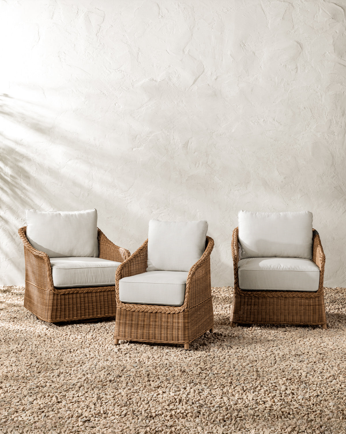 Three Haviland Outdoor Dining Chairs with Perennials Taupe Stripe cushions are set on a pebble-covered ground against a textured white wall, creating a relaxed and natural outdoor seating area by McGee & Co.