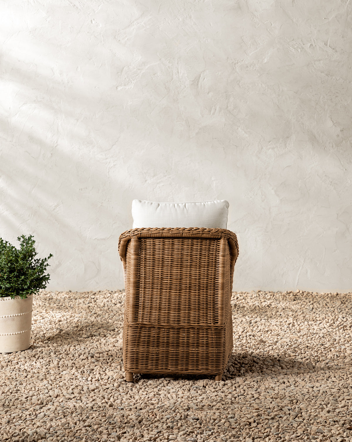 The Haviland Outdoor Dining Chair with a white cushion sits on a pebble-covered floor. To the left is a potted green plant, all set against a textured, light-colored wall in the background, by McGee & Co.