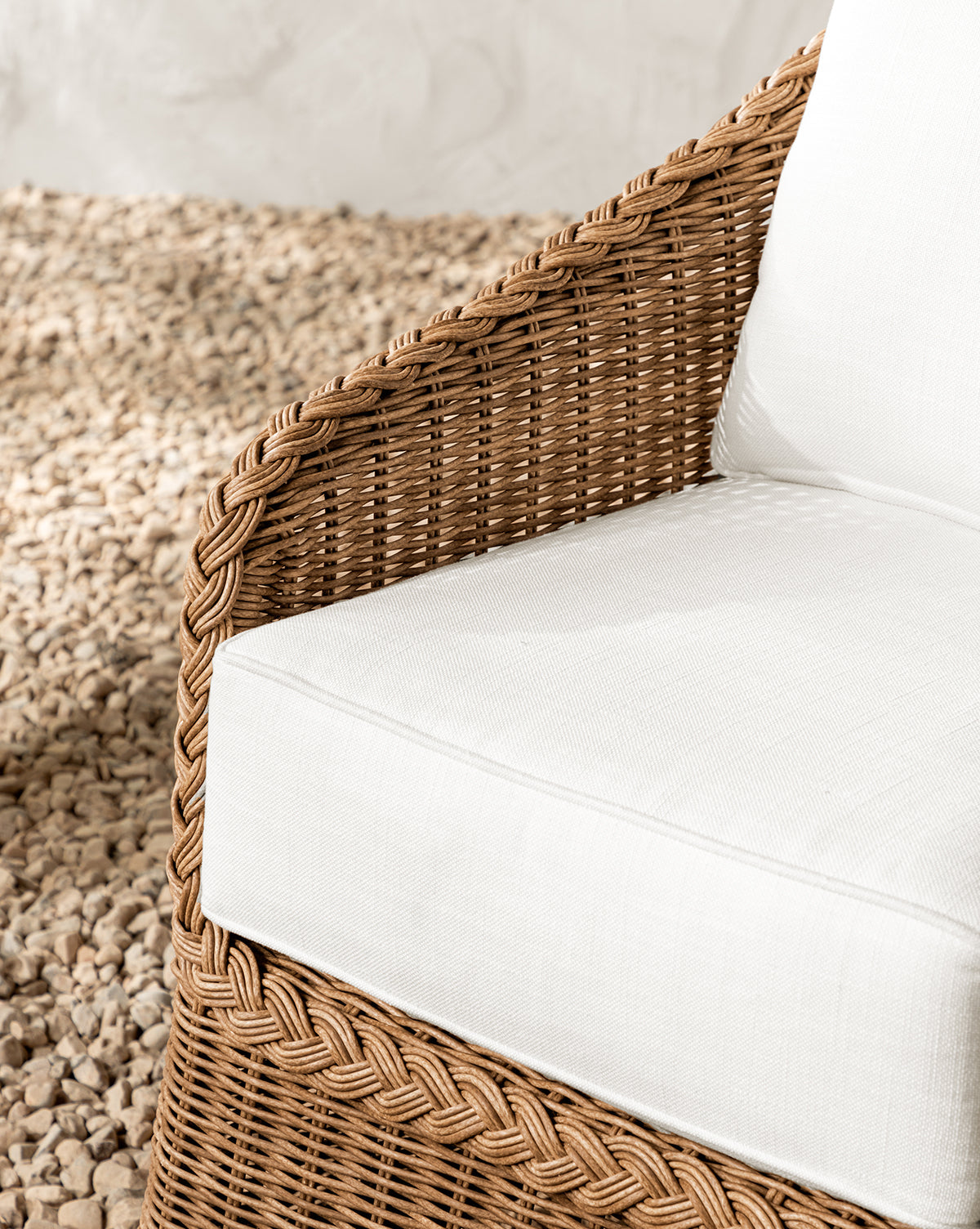 Close-up of the Haviland Outdoor Dining Chair, weather-resistant with a white cushion and pillow, set on light stones. Features include a braided wicker armrest and textured backrest, making it ideal for stylish outdoor dining, by McGee & Co.