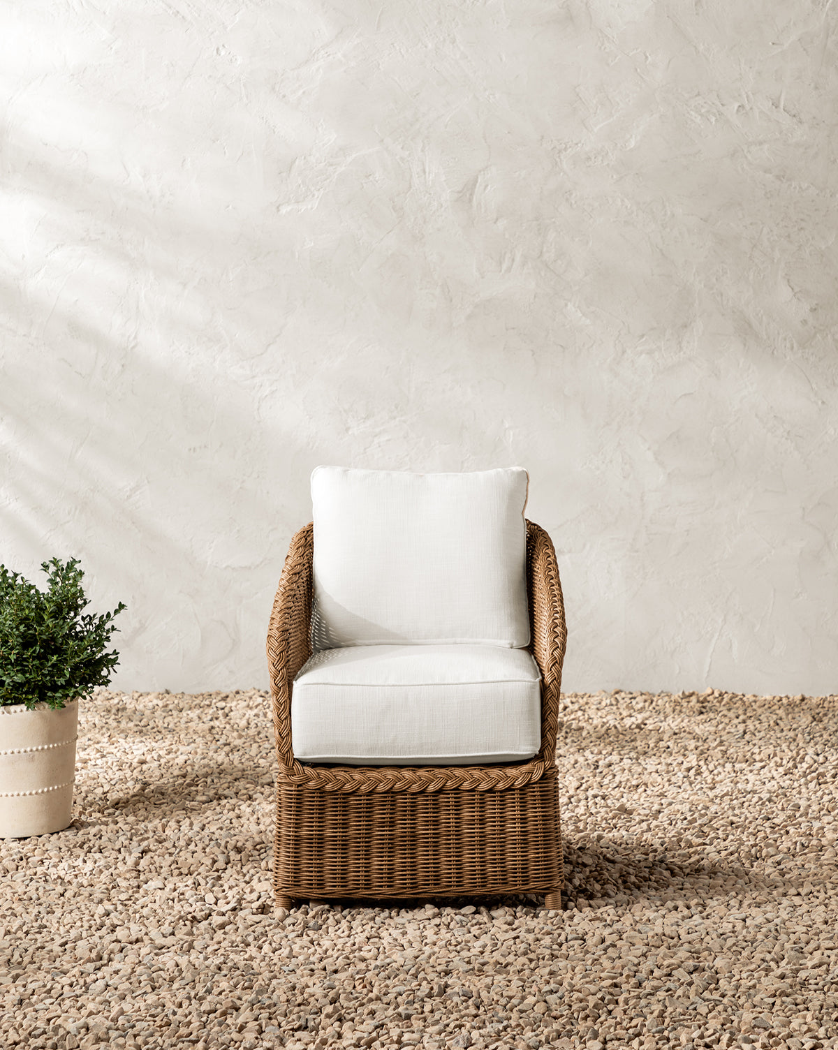 The Haviland Outdoor Dining Chair, featuring weather-resistant wicker and white cushions, is placed on gravel beside a potted green plant against a textured off-white wall, by McGee & Co.