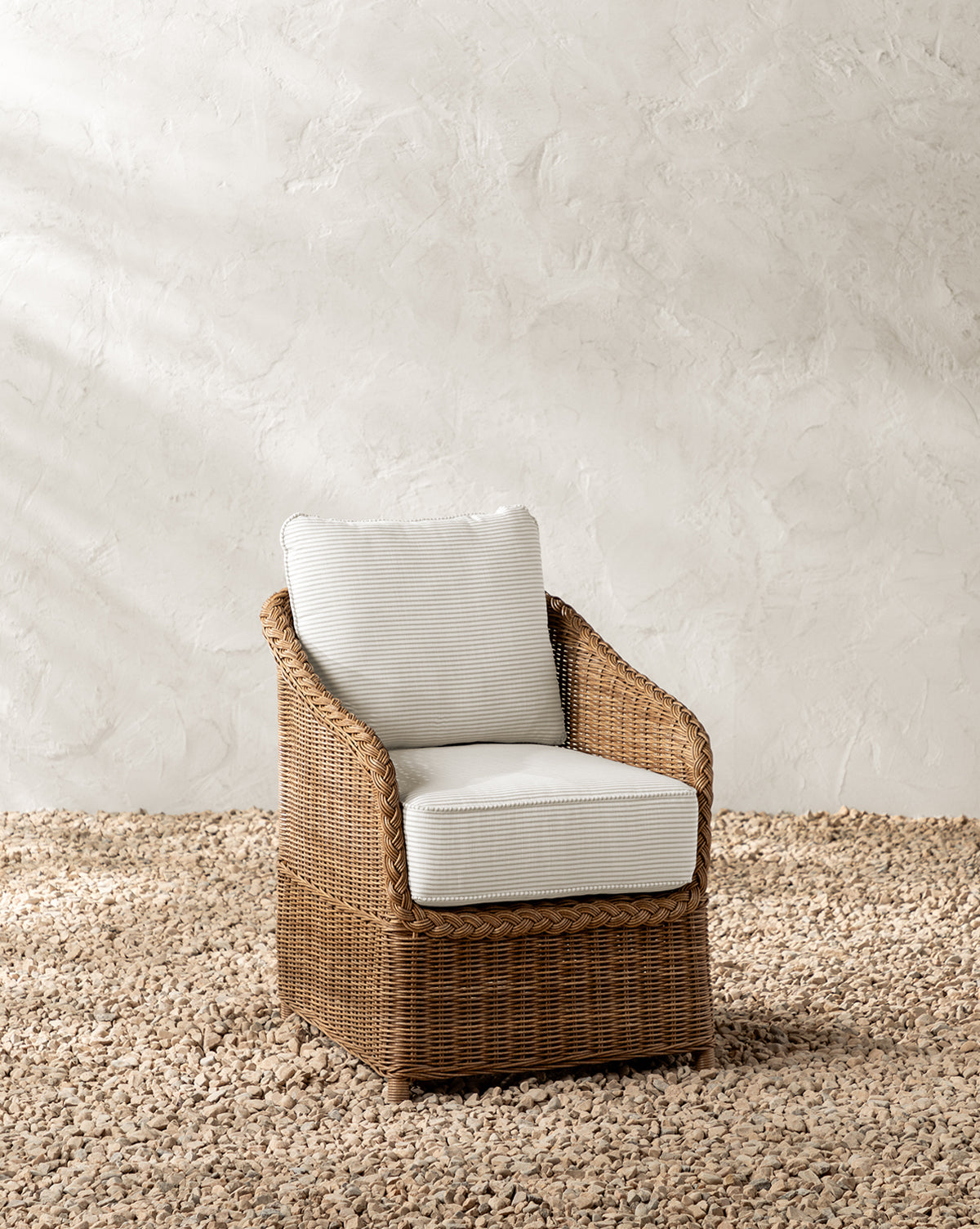 The Haviland Outdoor Dining Chair with Perennials Taupe Stripe sits on small, light pebbles against a textured off-white wall, featuring weather-resistant wicker and white cushions, by McGee & Co.