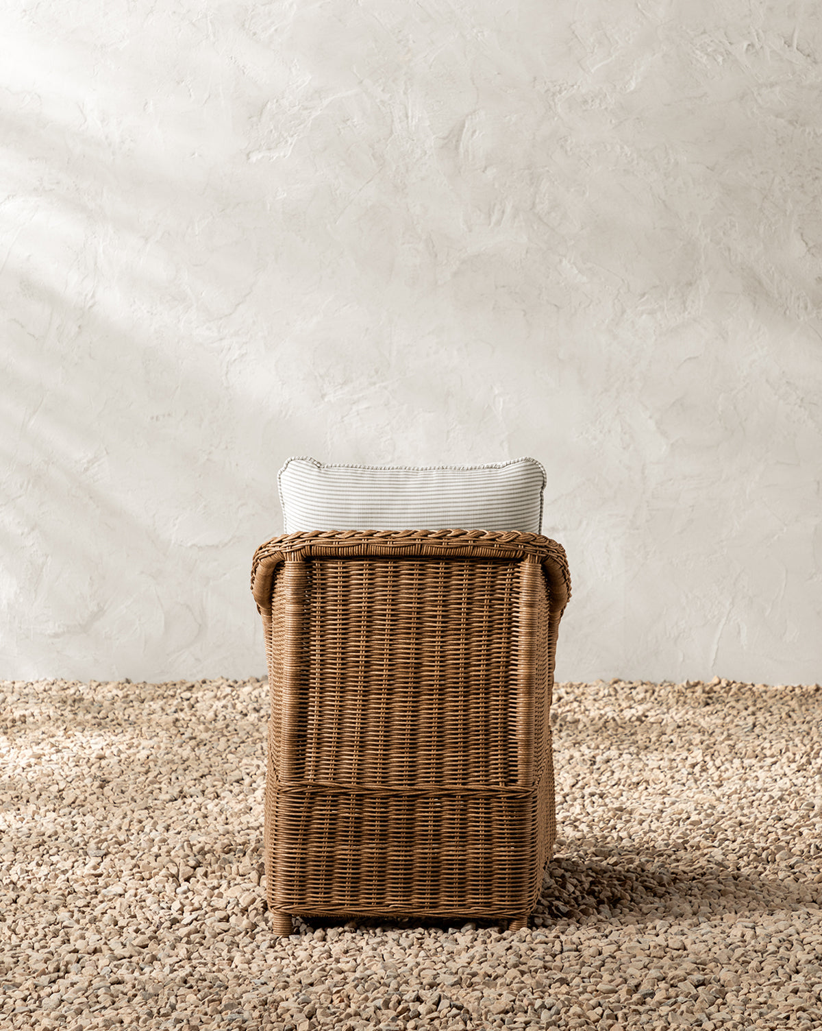 The Haviland Outdoor Dining Chair with Perennials Taupe Stripe, featuring a weather-resistant wicker frame and a light cushion, is placed on gravel against an off-white textured wall and shown from the back by McGee & Co.