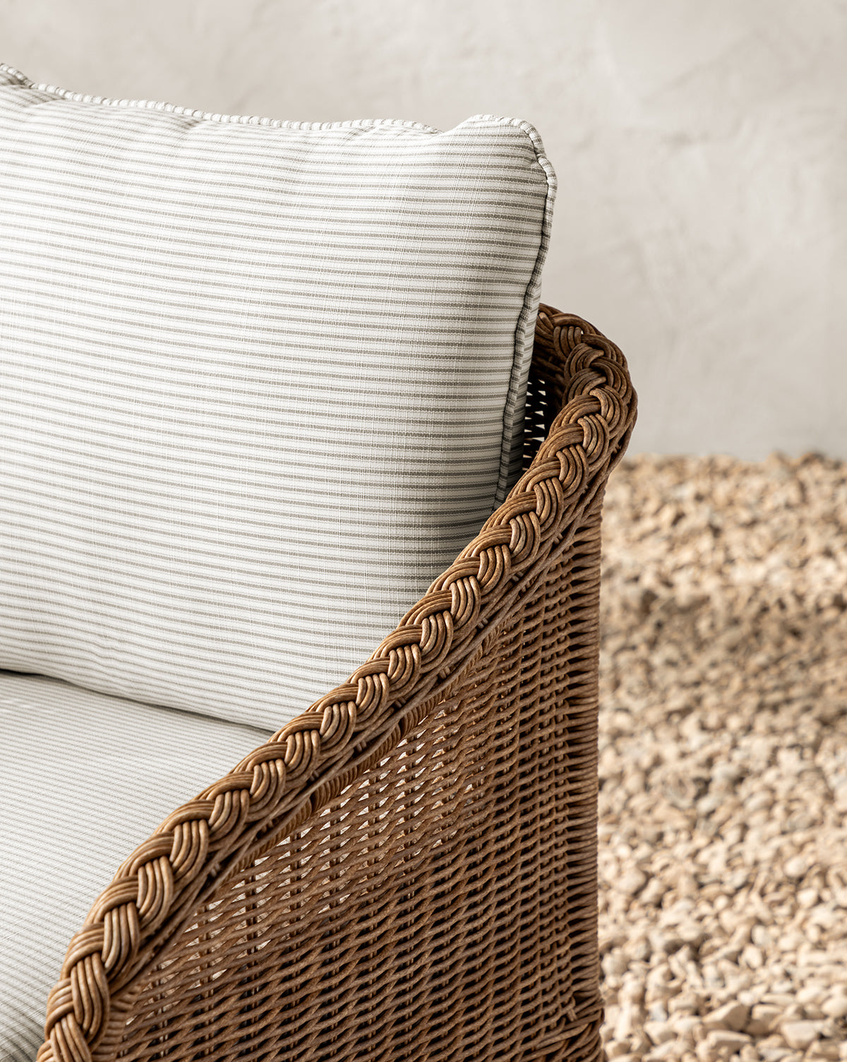 Close-up of the Haviland Outdoor Dining Chair with Perennials Taupe Stripe cushions, featuring a weather-resistant wicker frame, braided armrest detail, and set on light-colored stones before a textured wall, by McGee & Co.