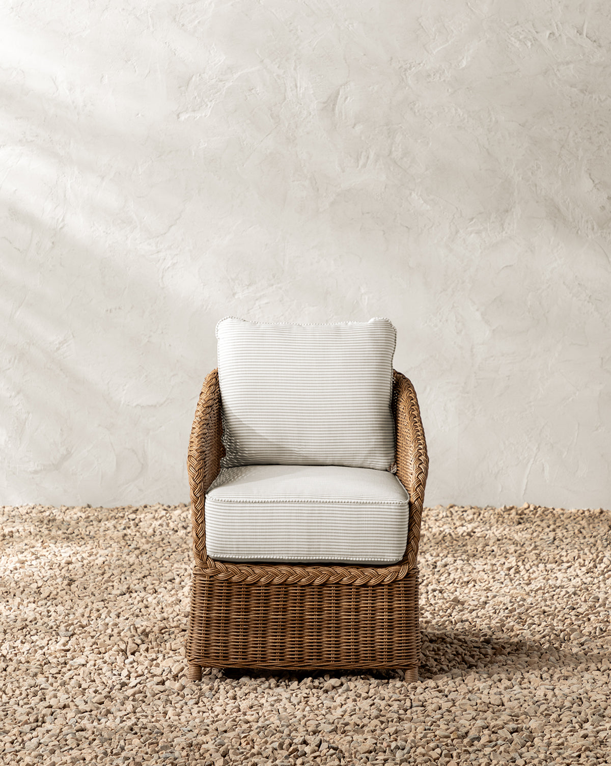 The Haviland Outdoor Dining Chair with Perennials Taupe Stripe and white cushions sits on a light gravel floor by a textured off-white wall, as sunlight casts soft shadows across the peaceful setting, by McGee & Co.