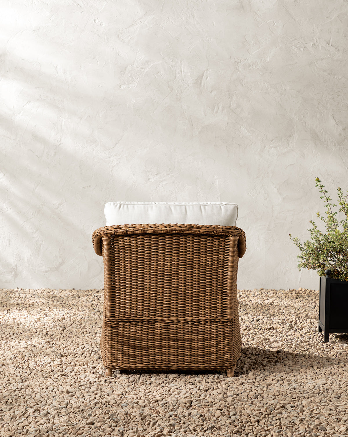 The Haviland Outdoor Lounge Chair with a white cushion sits on gravel before a textured light-colored wall, while a black planter with green foliage beside it brings natural charm to the scene, by McGee & Co.