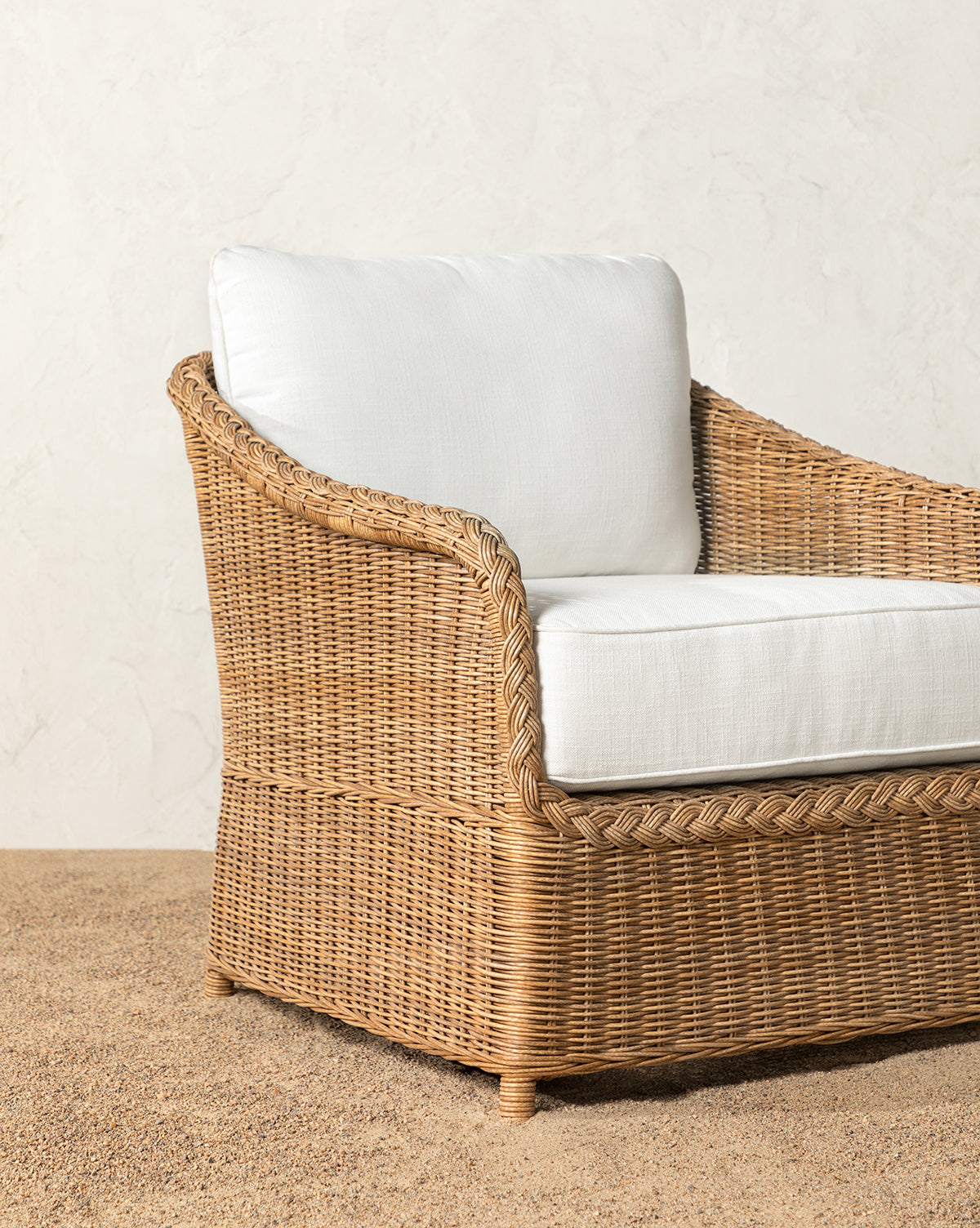 The Haviland Outdoor Lounge Chair, featuring a white cushion and weather-resistant wicker, is set on gravel before a textured white wall, accompanied by a potted plant on the right, by McGee & Co.
