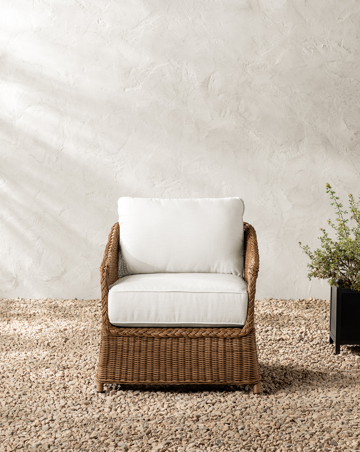 The Haviland Outdoor Lounge Chair with weather-resistant wicker and white cushions sits on gravel before a textured, light wall. To its right, a black rectangular planter filled with green foliage completes the scene, by McGee & Co.