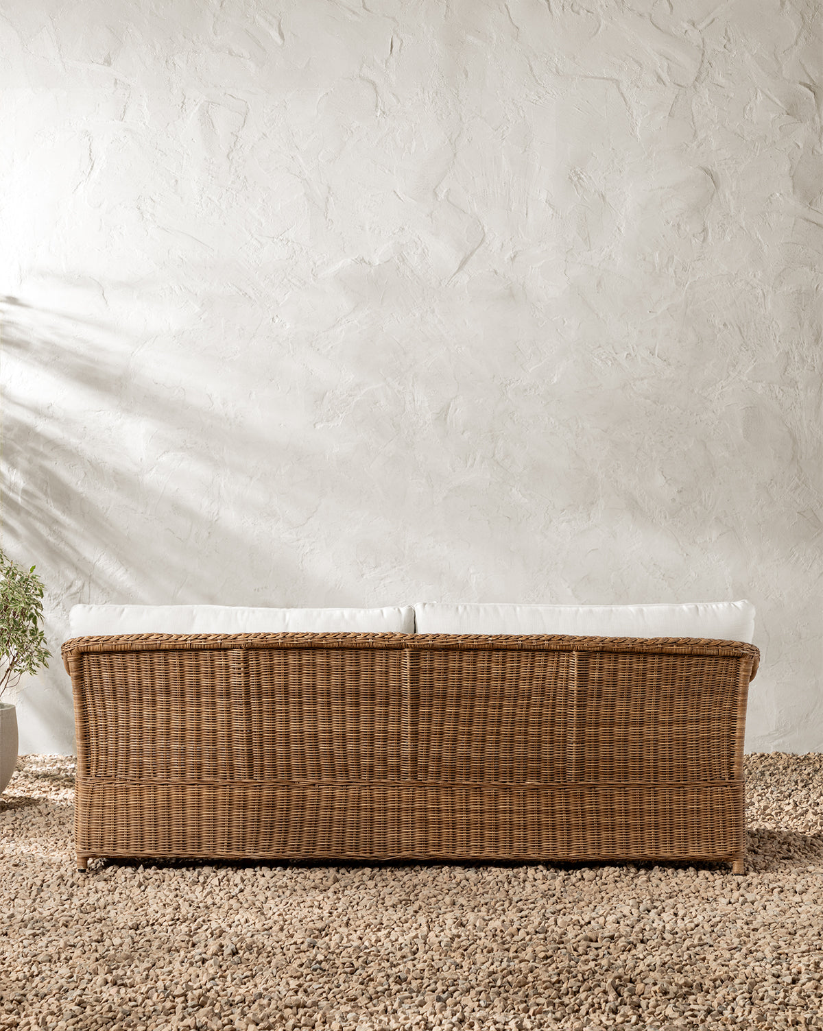 The Haviland Outdoor Sofa 88 with white cushions stands on a pebble floor before a textured white wall. A potted plant is on the left, while sunlight casts gentle shadows across the scene, by McGee & Co.