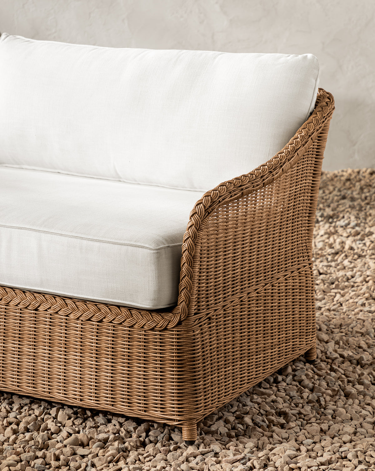 A close-up of the Haviland Outdoor Sofa 88, featuring weather-resistant wicker and white cushions, placed on small pebbles with a light-textured wall in the background, by McGee & Co.