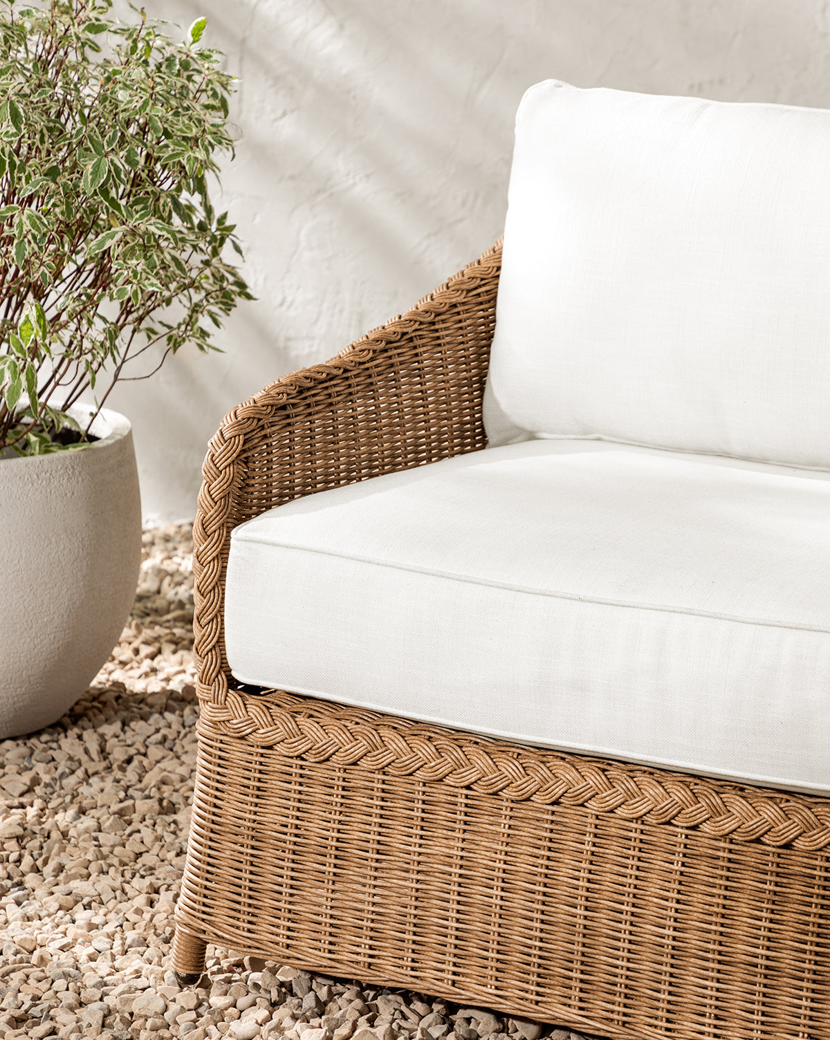The Haviland Outdoor Sofa 88 with weather-resistant wicker and white cushions sits on gravel by a large potted plant against a light wall, as sunlight casts soft shadows across the scene, by McGee & Co.