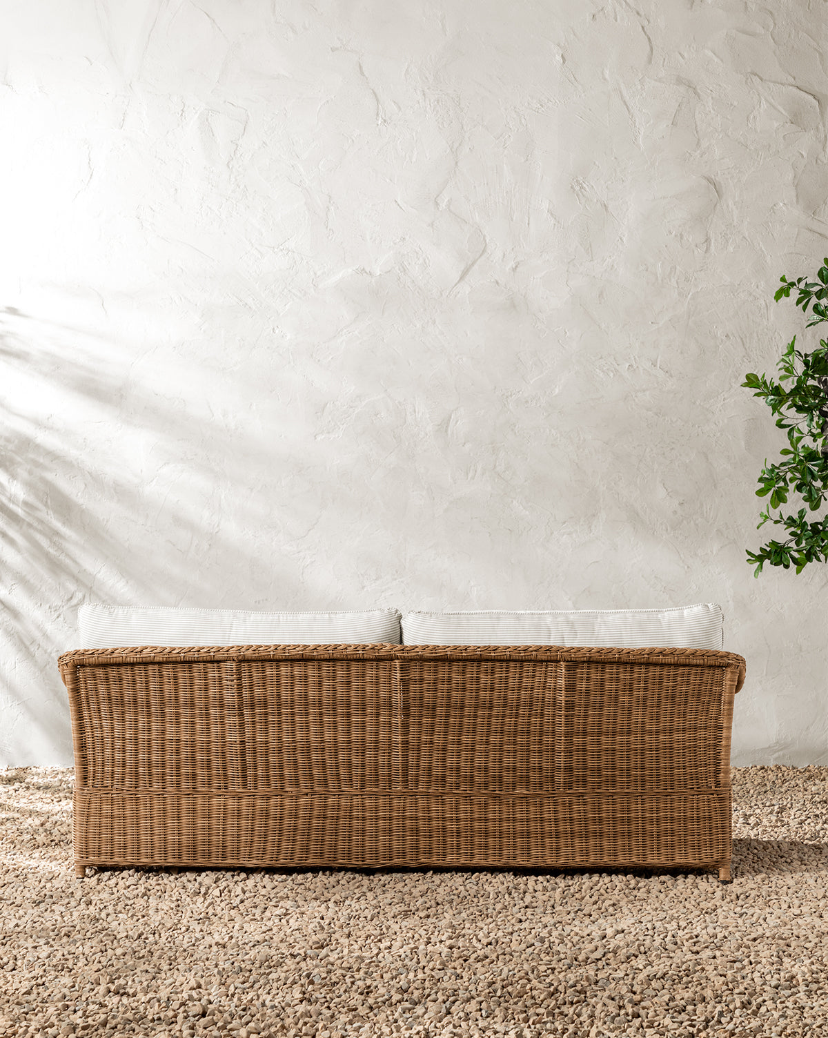 A Haviland Outdoor Sofa with Perennials Taupe Stripe 88 sits on a pebble floor against a textured white wall, complemented by a leafy green plant—a stylish, weather-resistant outdoor sofa perfect for relaxing, by McGee & Co.