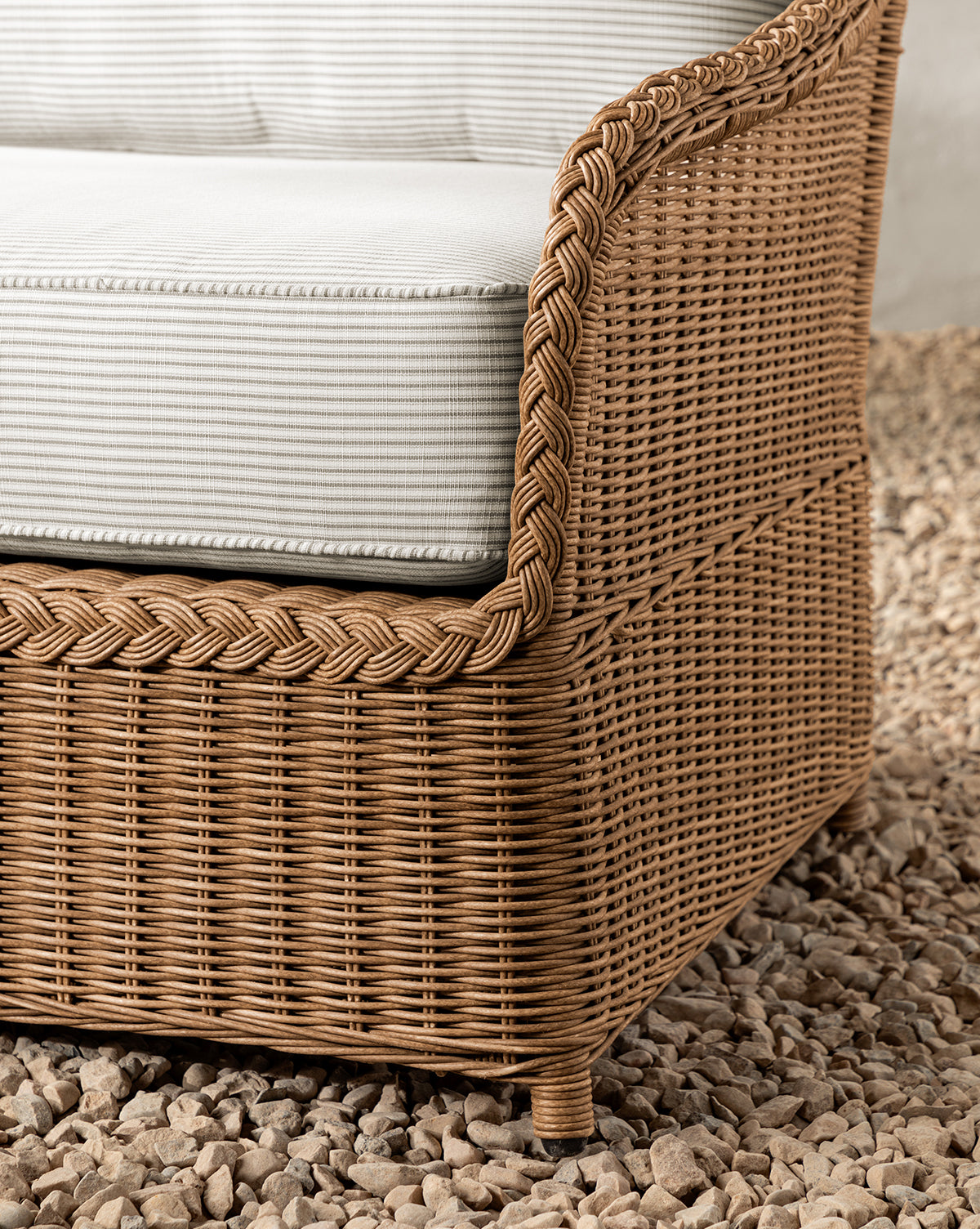Close-up of the Haviland Outdoor Sofa with Perennials Taupe Stripe 88, featuring a wicker frame with braided edge and light gray striped cushion, set atop small beige stones, by McGee & Co.