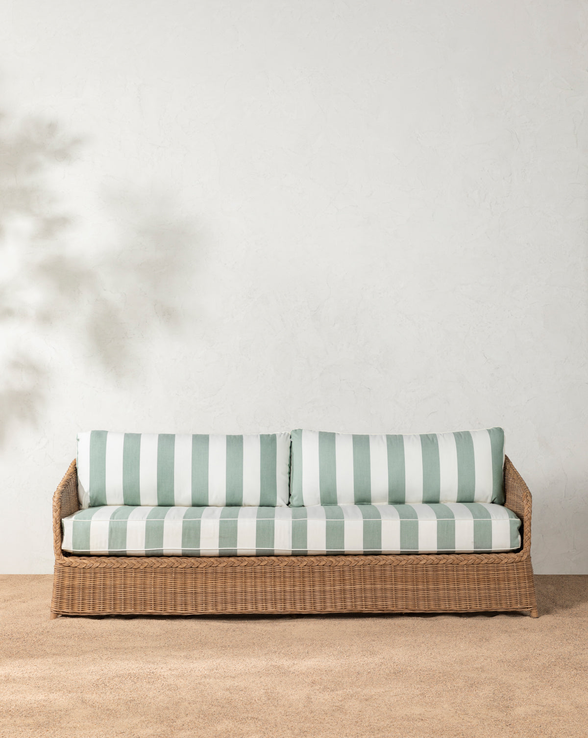 The Haviland Outdoor Sofa with Striped Cushions 88 rests on a gravel floor against a textured white wall, featuring light green and white stripes. A small potted plant sits to the right as leafy shadows fall gently across the scene, by McGee & Co.
