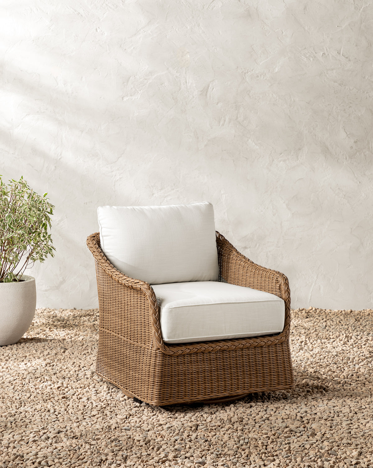The Haviland Outdoor Swivel Lounge Chair with white cushions sits on a pebble floor beside a potted green plant and a textured light wall, while sunlight casts gentle shadows across the serene outdoor scene, by McGee & Co.