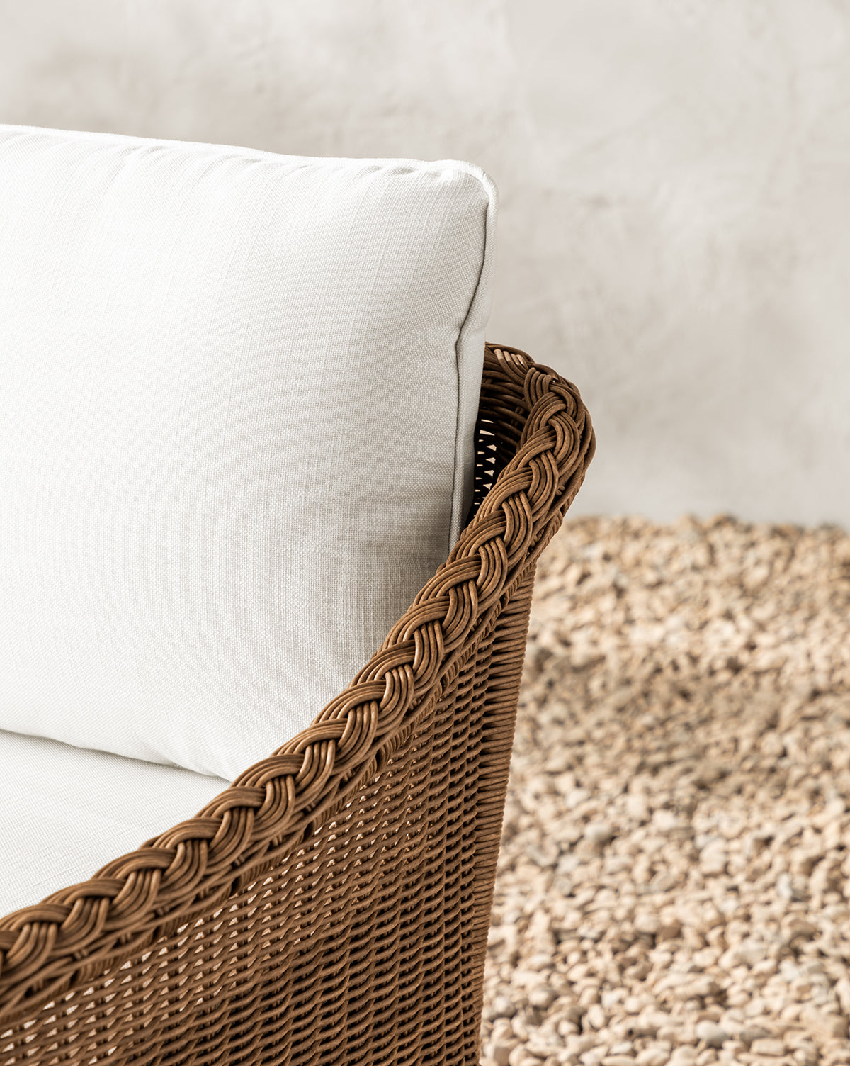 Close-up of the Haviland Outdoor Swivel Lounge Chair, featuring weather-resistant brown wicker and a white cushion, set on a pebble-textured surface with a light gray, slightly textured wall in the background, by McGee & Co.
