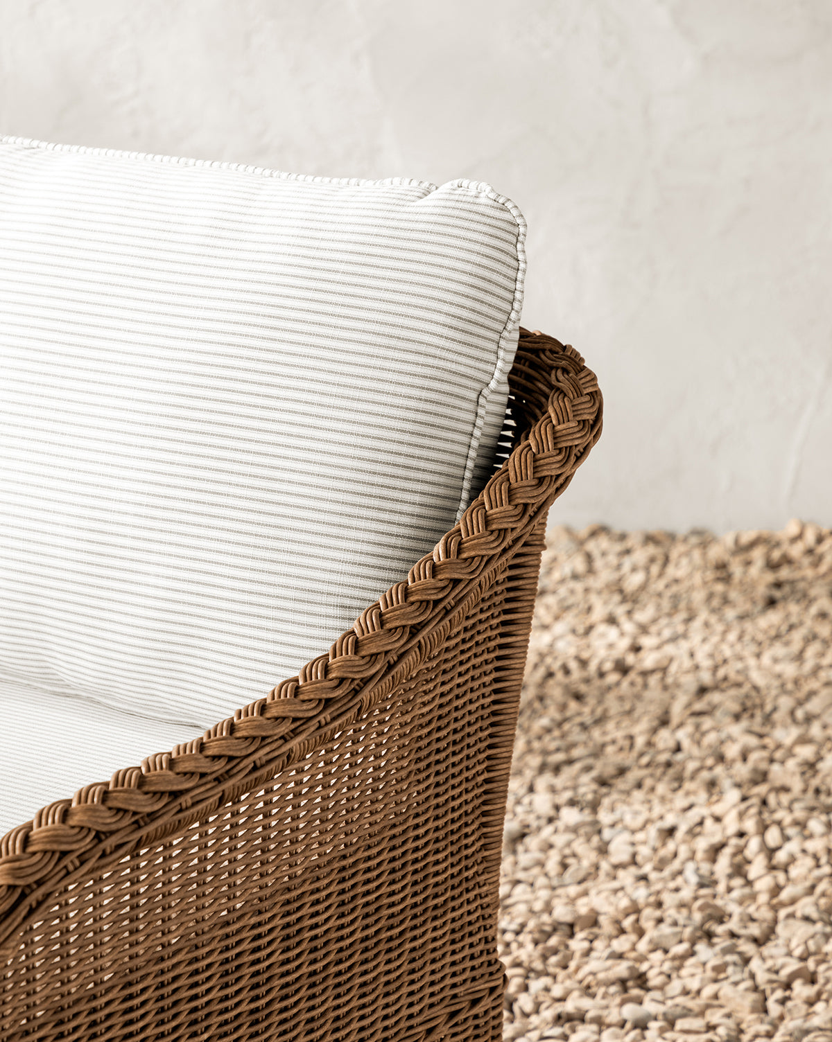 The Haviland Outdoor Swivel Lounge Chair features a wicker frame and a white striped cushion, set on a light pebble floor by an off-white textured wall—ideal as a stylish, weather-resistant outdoor chair, by McGee & Co.