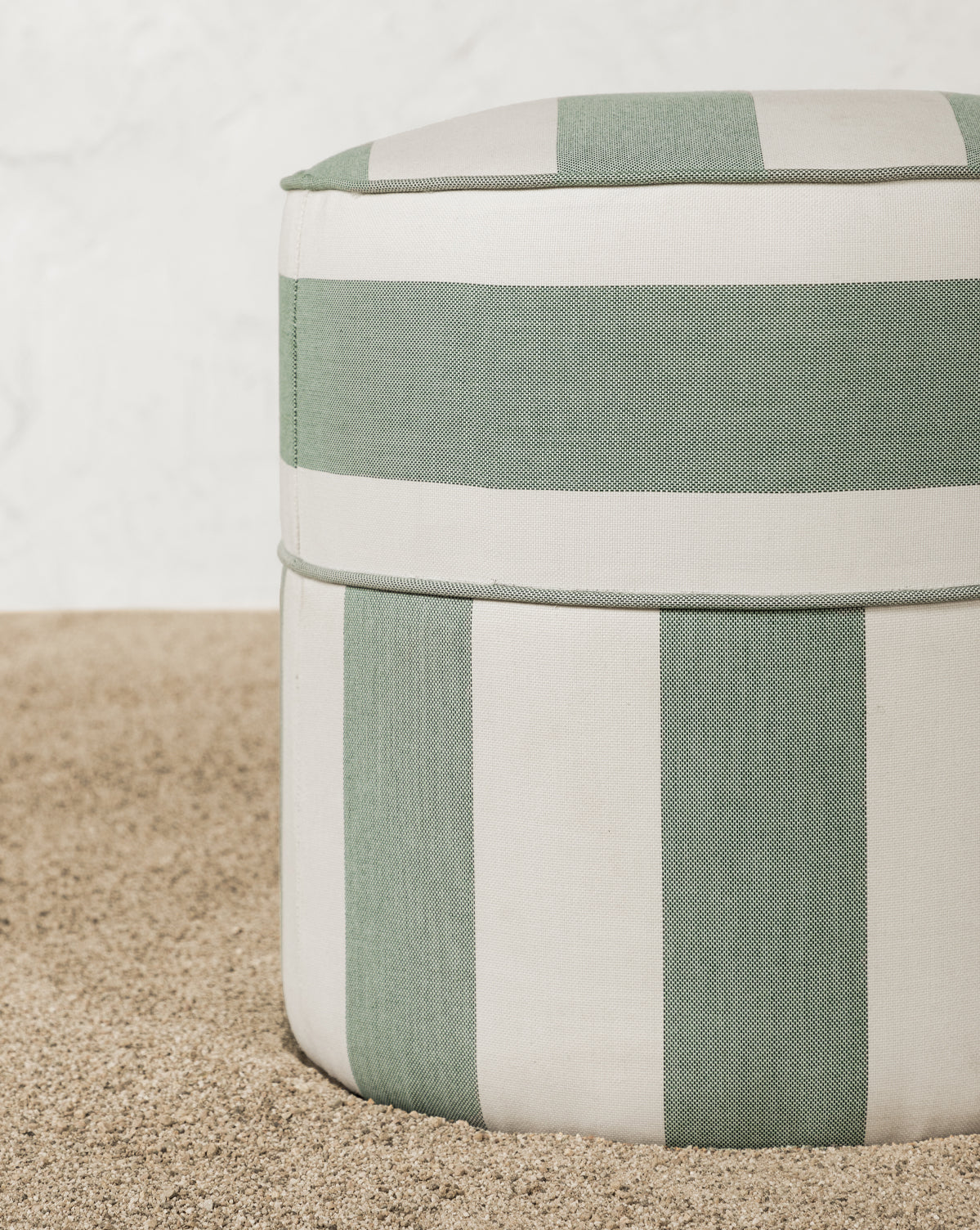 The Makerspalm Haviland Striped Ottoman features light green and white vertical stripes, sits on a pebble floor by a textured white wall, and is weather-resistant—perfect as a stylish outdoor accent. McGee & Co.