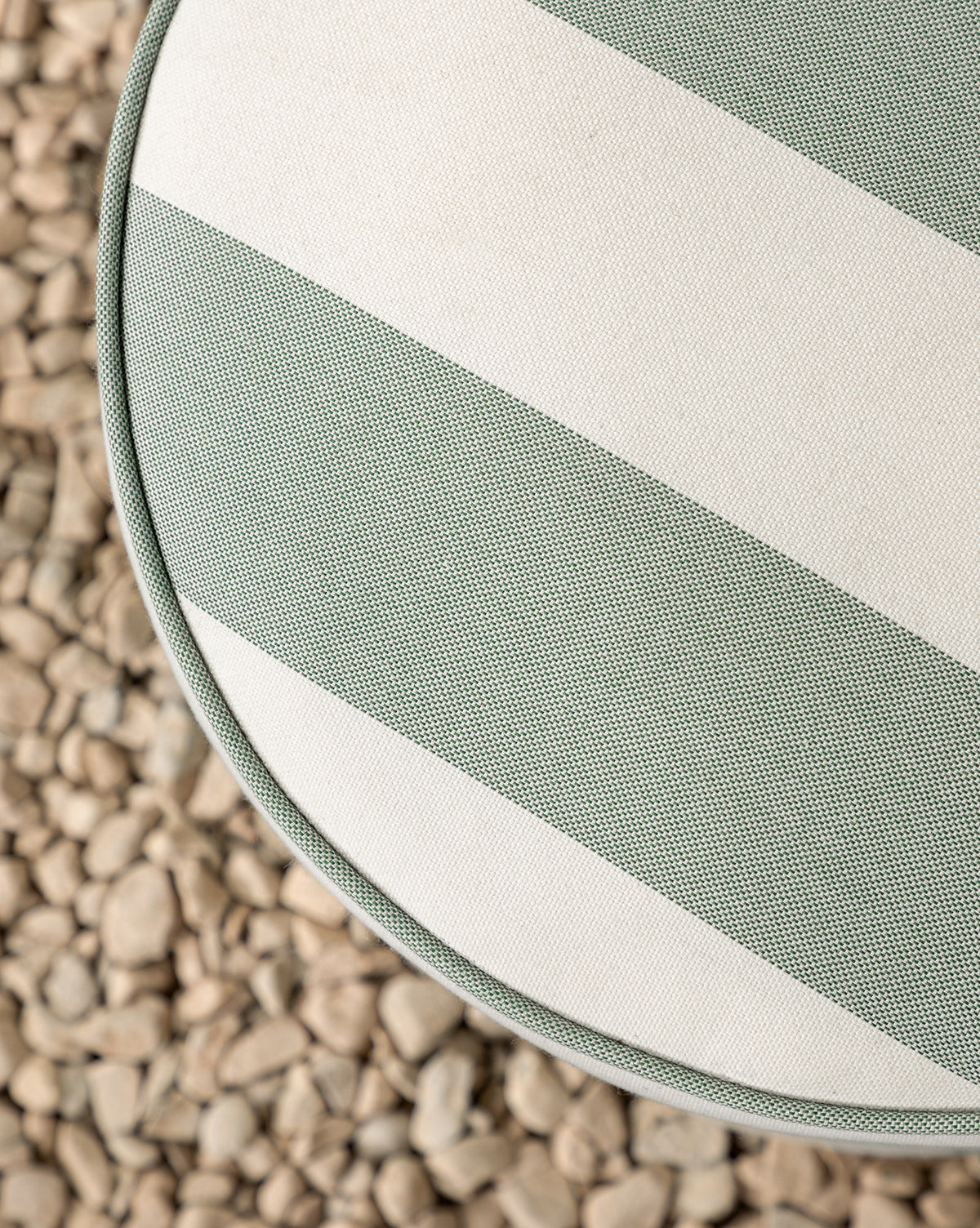 The Makerspalm Haviland Striped Ottoman, featuring wide green and white weather-resistant stripes, is shown from above on a bed of small, light-colored pebbles, with a partial view of its cushion and the textured surface. McGee & Co.