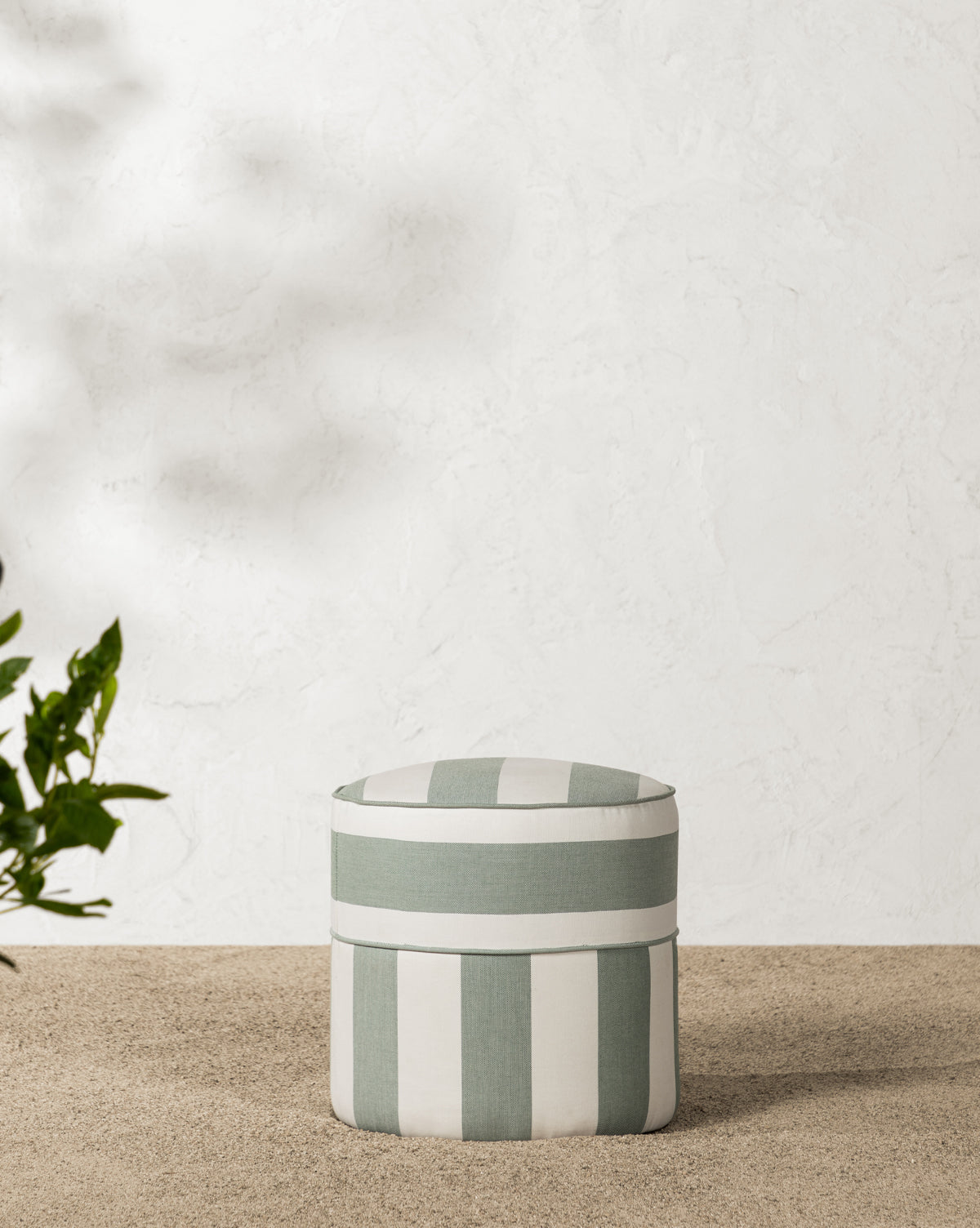 The Makerspalm Haviland Striped Ottoman in light green and white sits on a textured pebble floor by a white wall, with sunlight casting soft shadows that highlight this weather-resistant outdoor ottoman’s inviting style. McGee & Co.