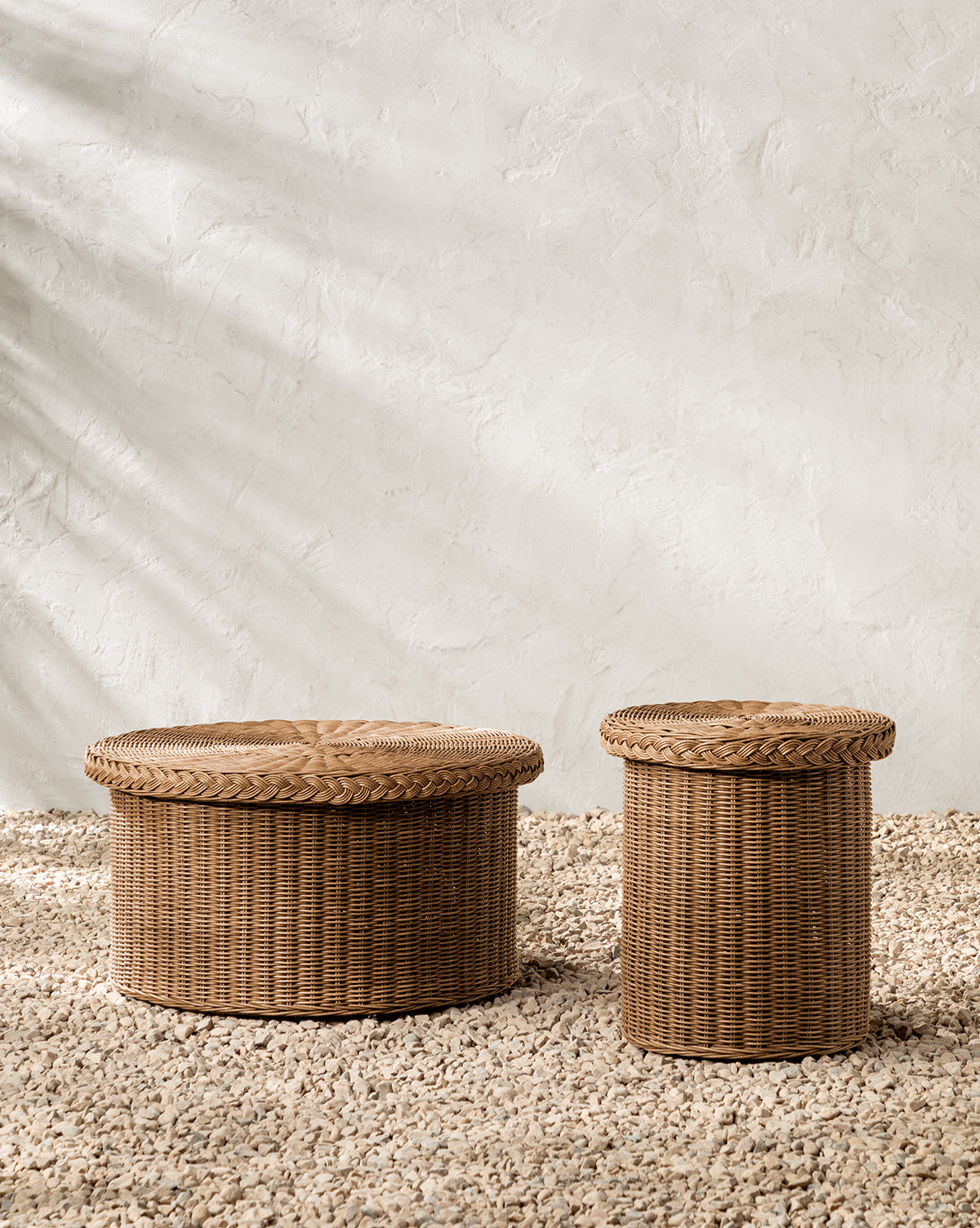 The Haviland Side Table, featuring two round wicker baskets with lids in varying sizes, sits on a pebble floor against a light stucco wall—ideal as weather-resistant accents or as a multifunctional outdoor side table, by McGee & Co.