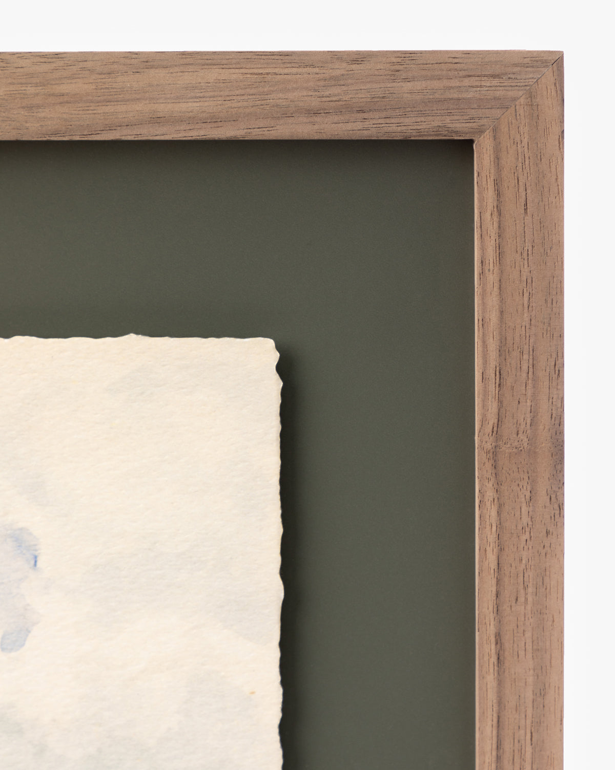 Close-up of the corner of a walnut frame with a dark green mat, showing Head in the Clouds—a textured, off-white print on paper inside the frame.