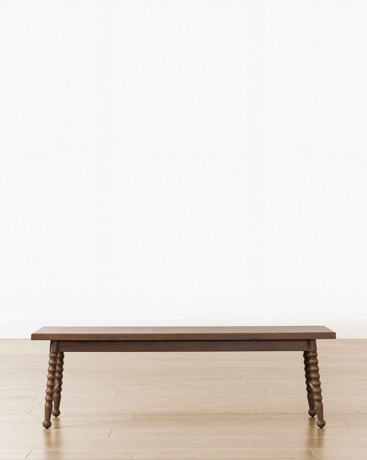 A Heathcliff Bench with turned legs sits on a light wood floor against a plain white wall, by McGee & Co.