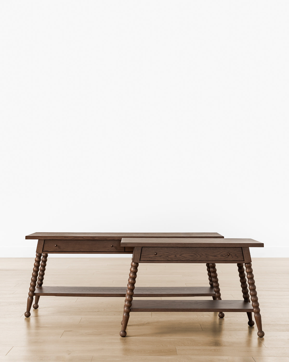 Two Heathcliff Console tables of varying heights feature turned bobbin legs, a lower shelf, and a single drawer each. Their subtle walnut oak finish highlights classic charm against a plain white background and light wood floor, by McGee & Co.