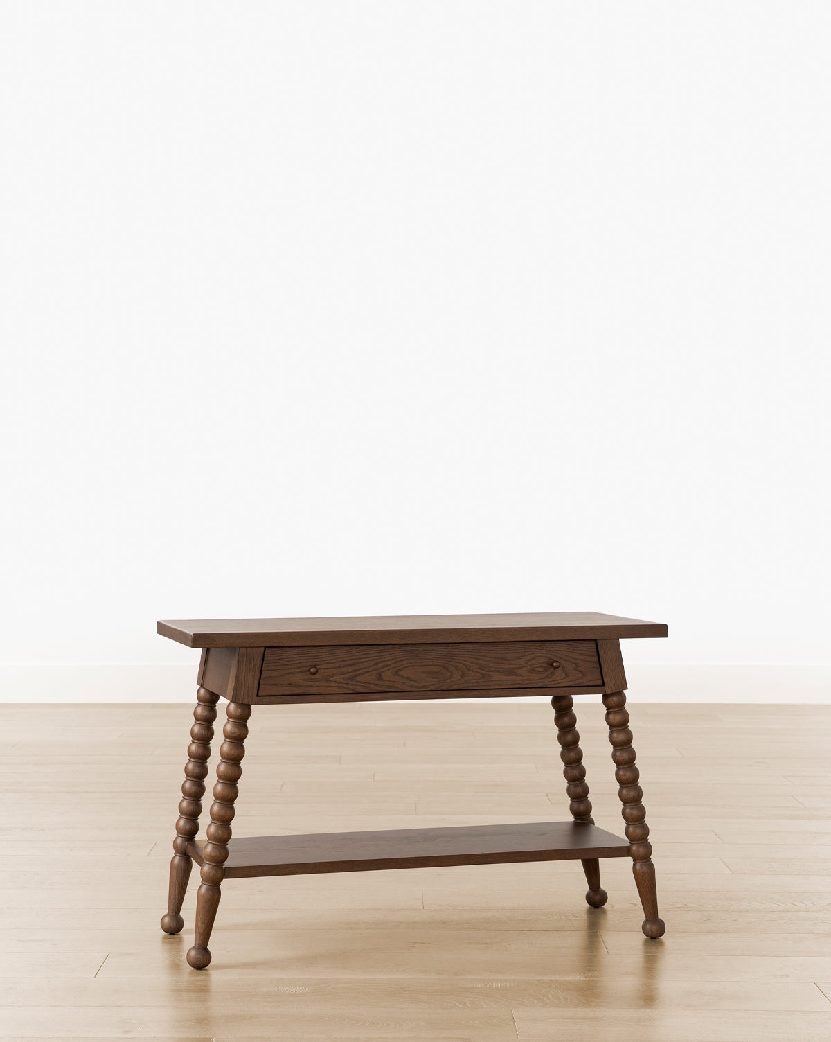 The Heathcliff Console is a wooden side table featuring turned bobbin-style legs, a single front drawer, and a lower shelf, shown on a light wood floor against a plain white background, by McGee & Co.