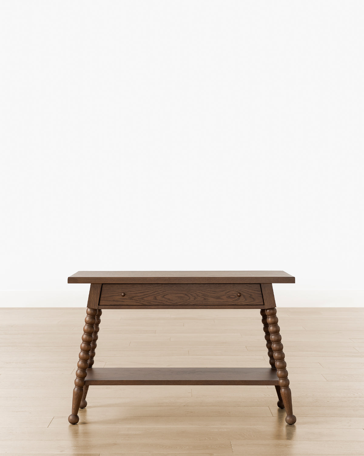 The Heathcliff Console, a wooden side table with a walnut oak finish, single drawer, lower shelf, and spiral-turned legs, stands on a light wood floor against a plain white background, by McGee & Co.
