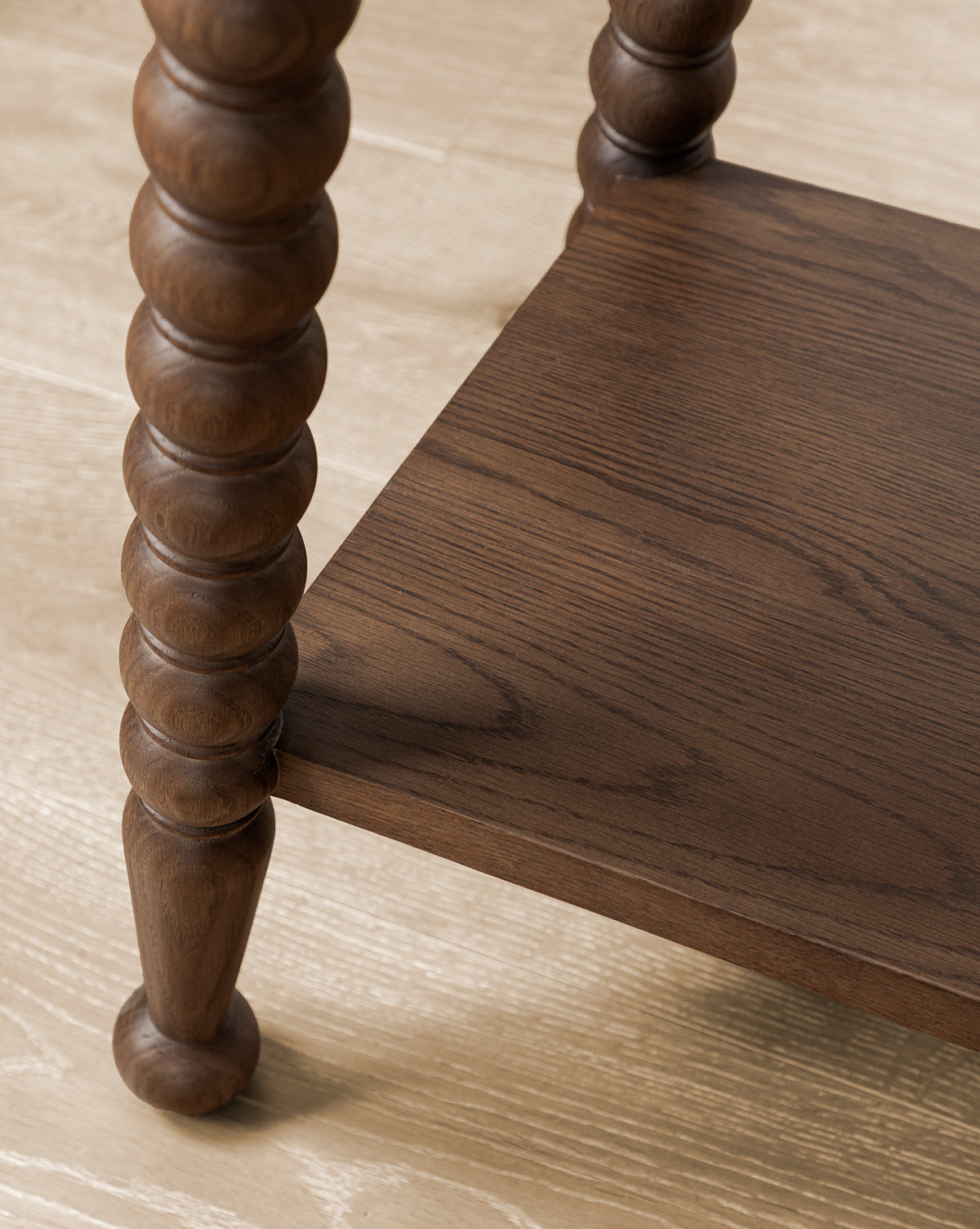 A close-up view of the lower shelf and leg of the Heathcliff Console, featuring round carved bobbin detailing and a walnut oak finish, set on a light wood floor, by McGee & Co.