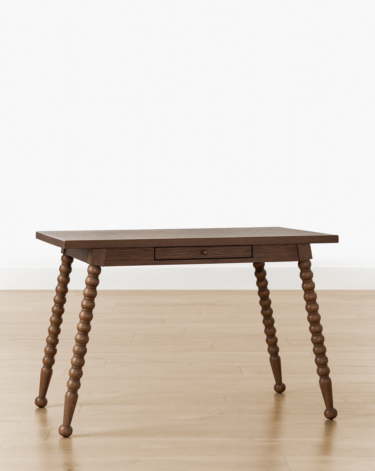 The Heathcliff Desk features a bobbin-style design with a central drawer and four spindle-turned legs, shown on a light wood floor against a plain white background by McGee & Co.