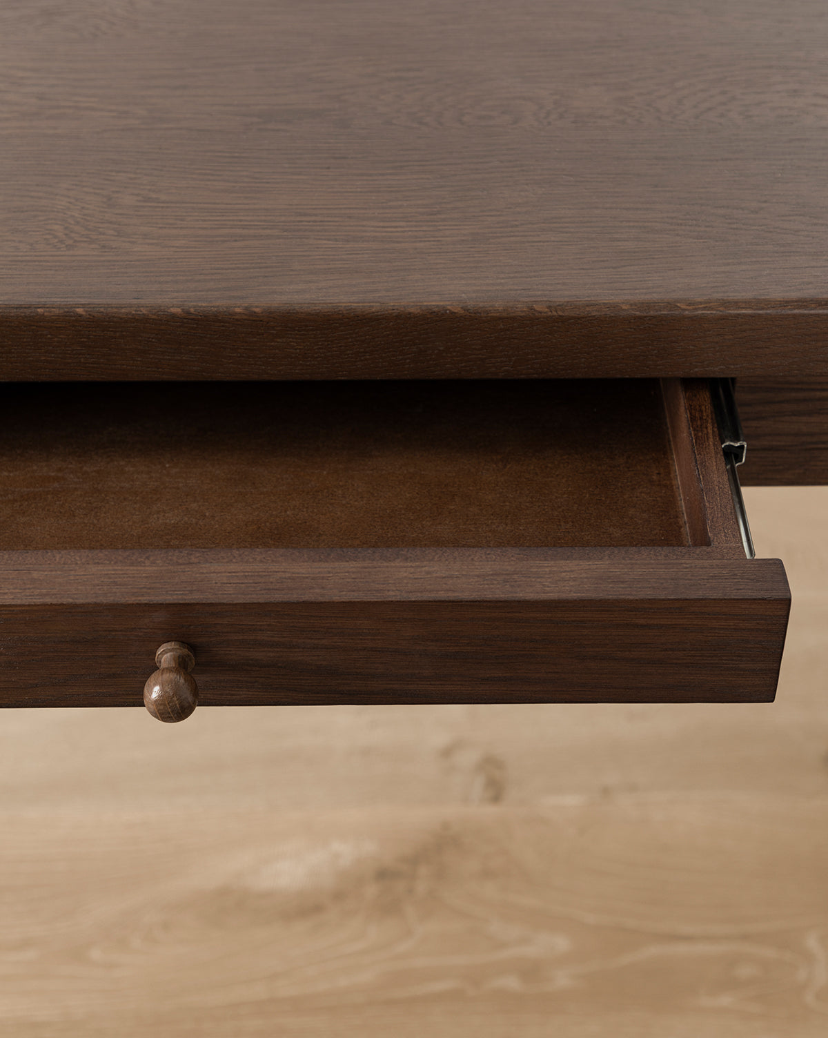 A close-up of the Heathcliff Desks partially open wooden drawer with a round knob, shown against a light wood floor background by McGee & Co.