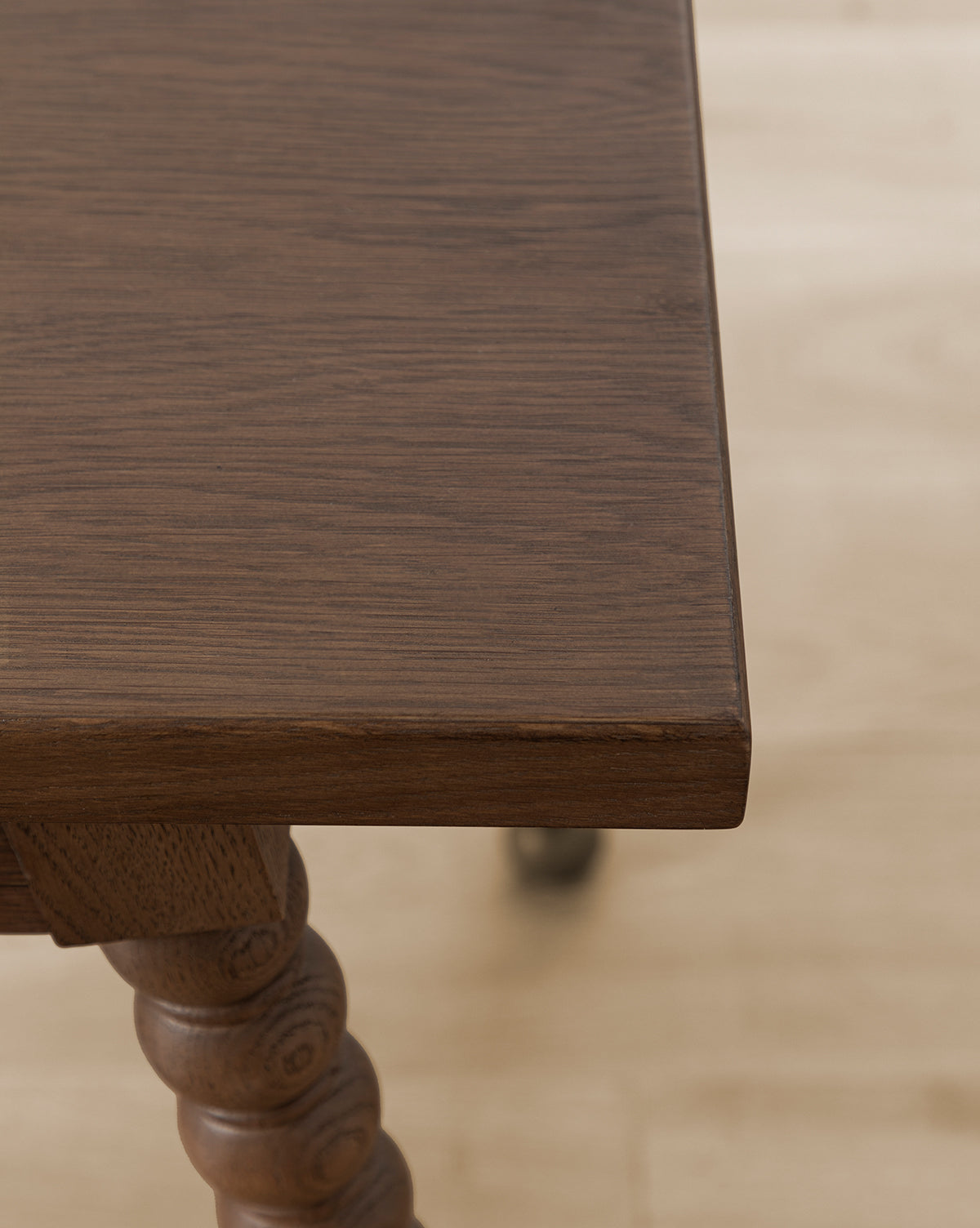 A close-up of the Heathcliff Desk’s corner shows a smooth, dark brown walnut oak surface and a carved spiral bobbin-style leg, set against a light-colored floor, by McGee & Co.
