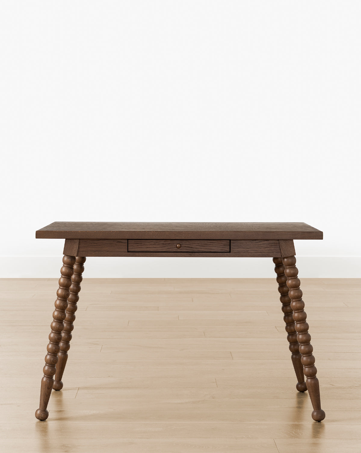 The Heathcliff Desk features a walnut oak finish, single center drawer, and four bobbin-style spindle legs. It stands on a light wood floor against a plain white wall, by McGee & Co.