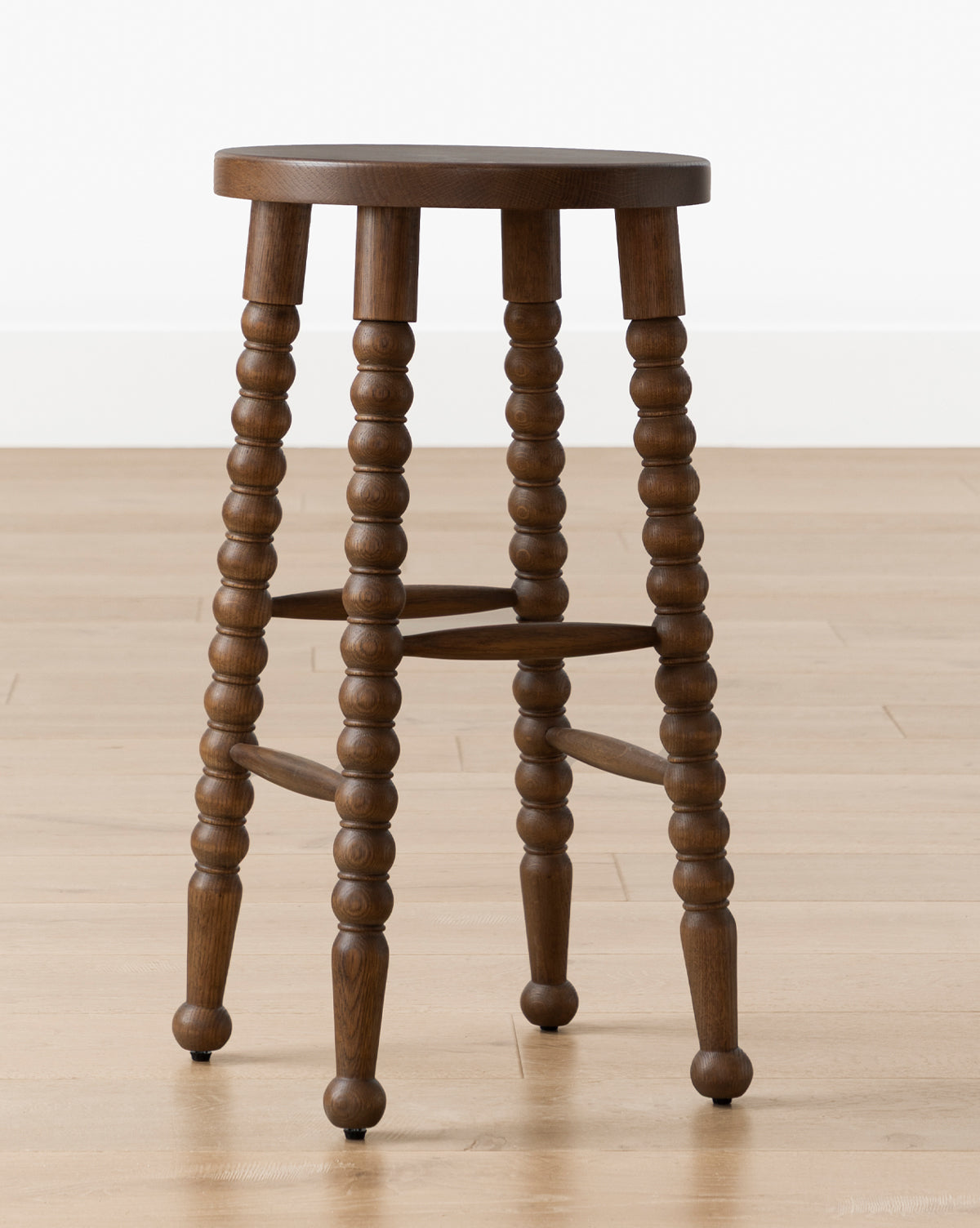 A Makerspalm Heathcliff Stool with a round wooden seat and four spindle-style legs in walnut oak finish, sits on a light wood floor against a white wall, McGee & Co.
