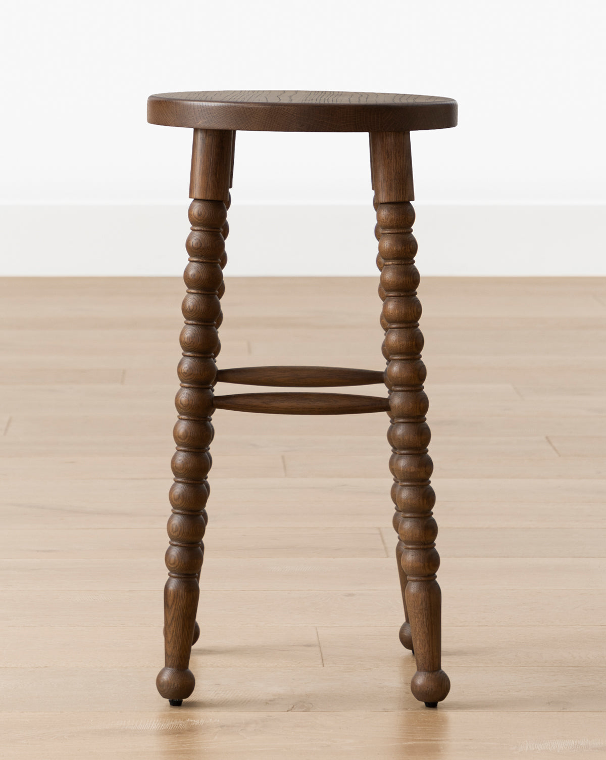 The Makerspalm Heathcliff Stool, featuring a walnut oak finish, turned spindle legs, and a round seat, is placed on a light wood floor against a white wall. McGee & Co.